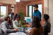 Team meeting in a modern office, with a man presenting to seated colleagues around a desk.