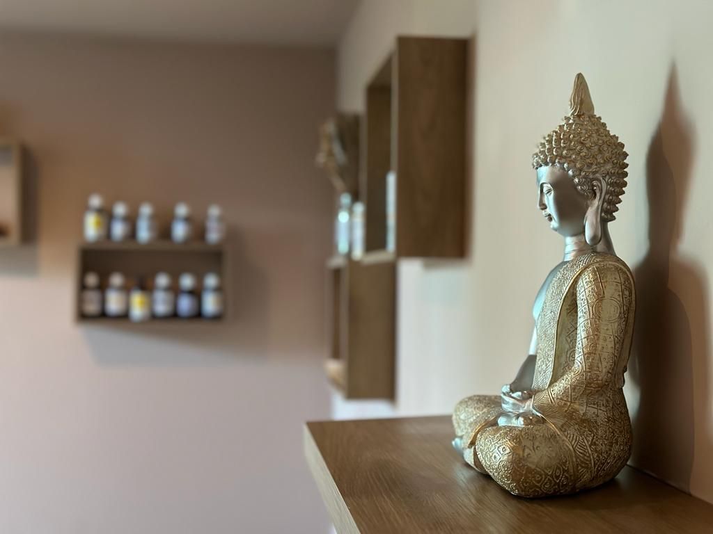 Golden Buddha statue on a wooden shelf in a softly lit room with small bottles on wall shelves.