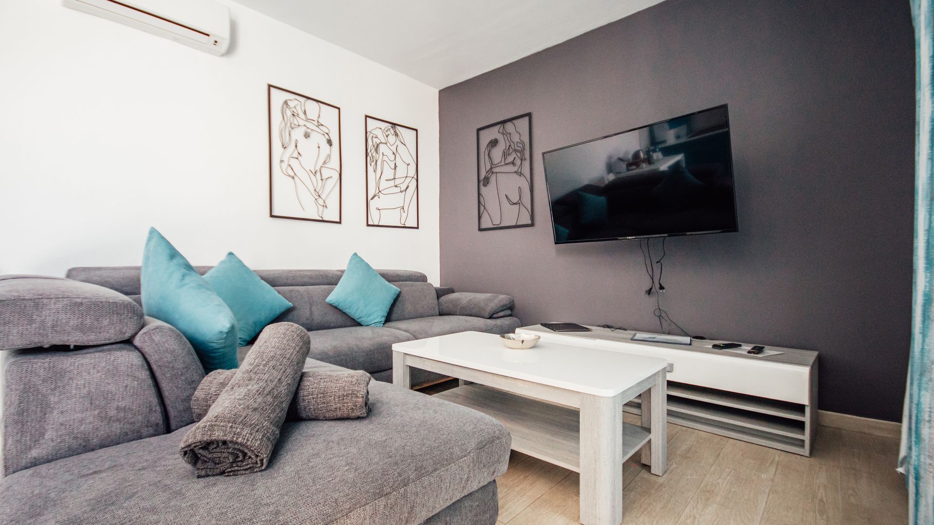 Modern gray living room with sectional sofa, teal pillows, wall-mounted TV, and white coffee table.