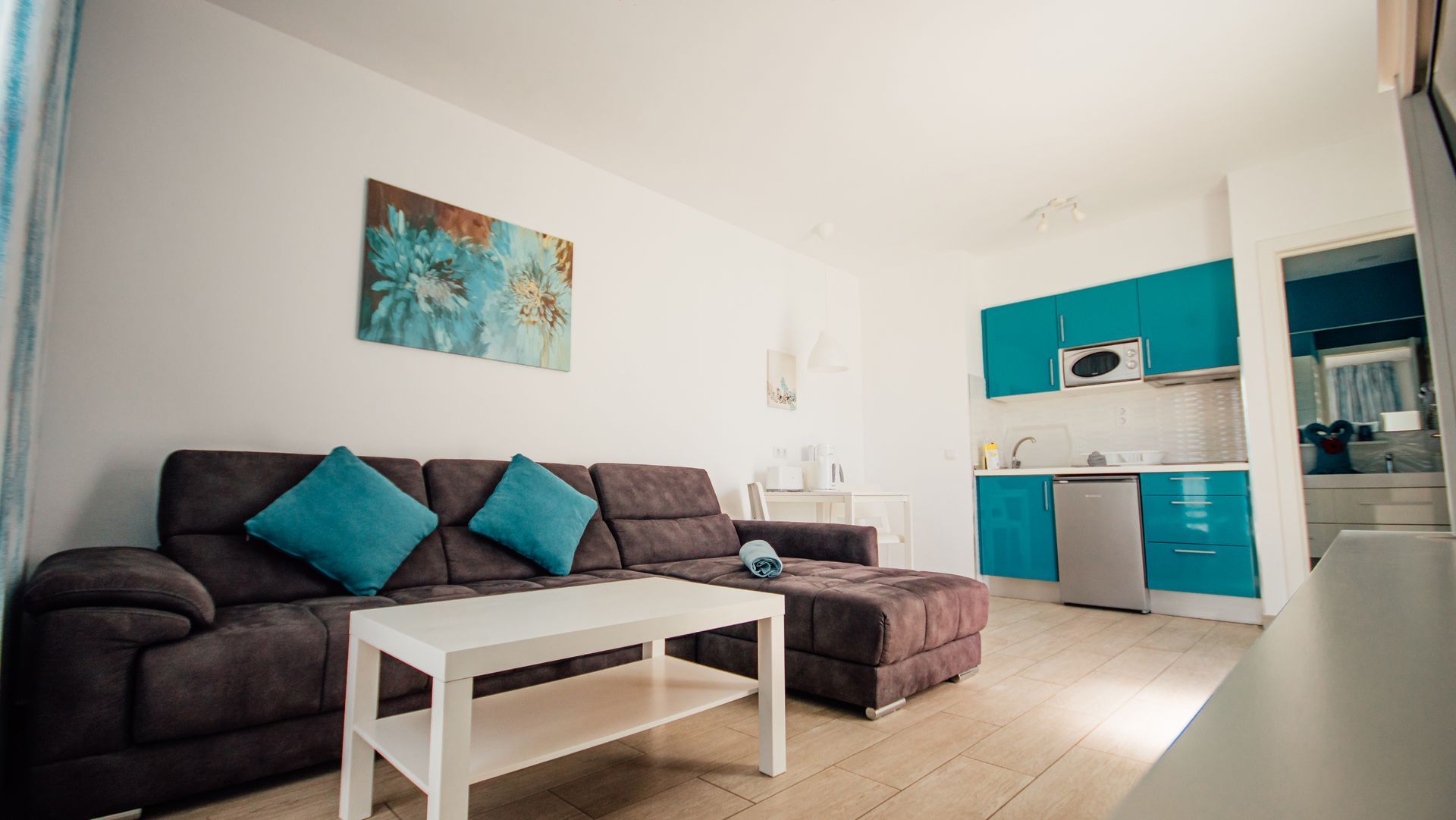 Modern living room with dark sofa, turquoise cushions, white coffee table, and teal kitchenette.