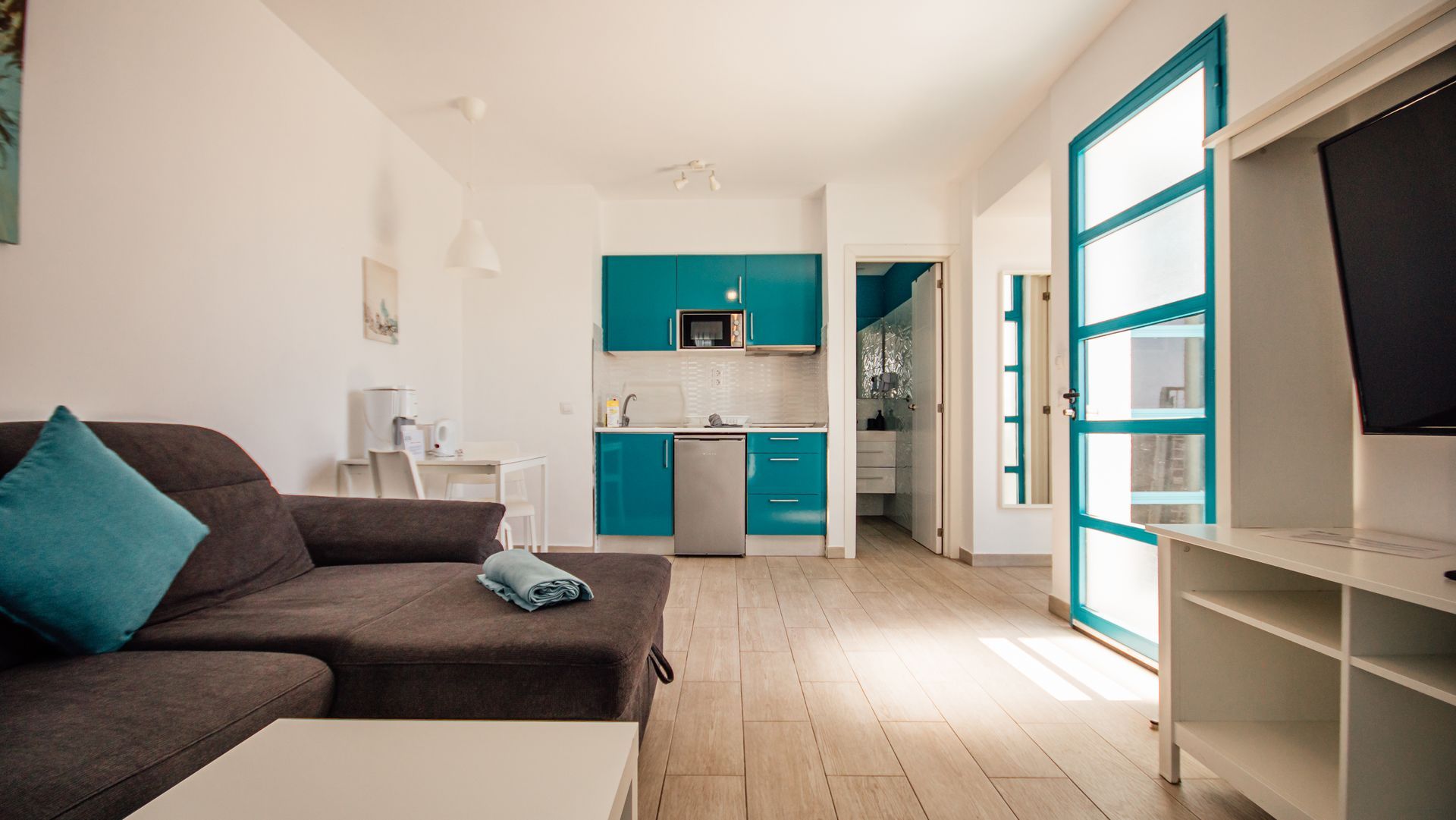 Bright living room with gray sofa, teal accents, and a compact kitchen in the background