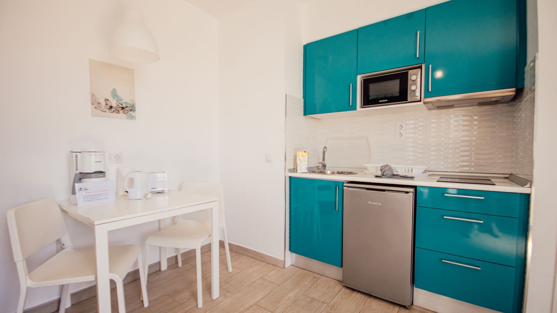 Compact kitchen with teal cabinets, stainless appliances, and a small white dining table by the wall