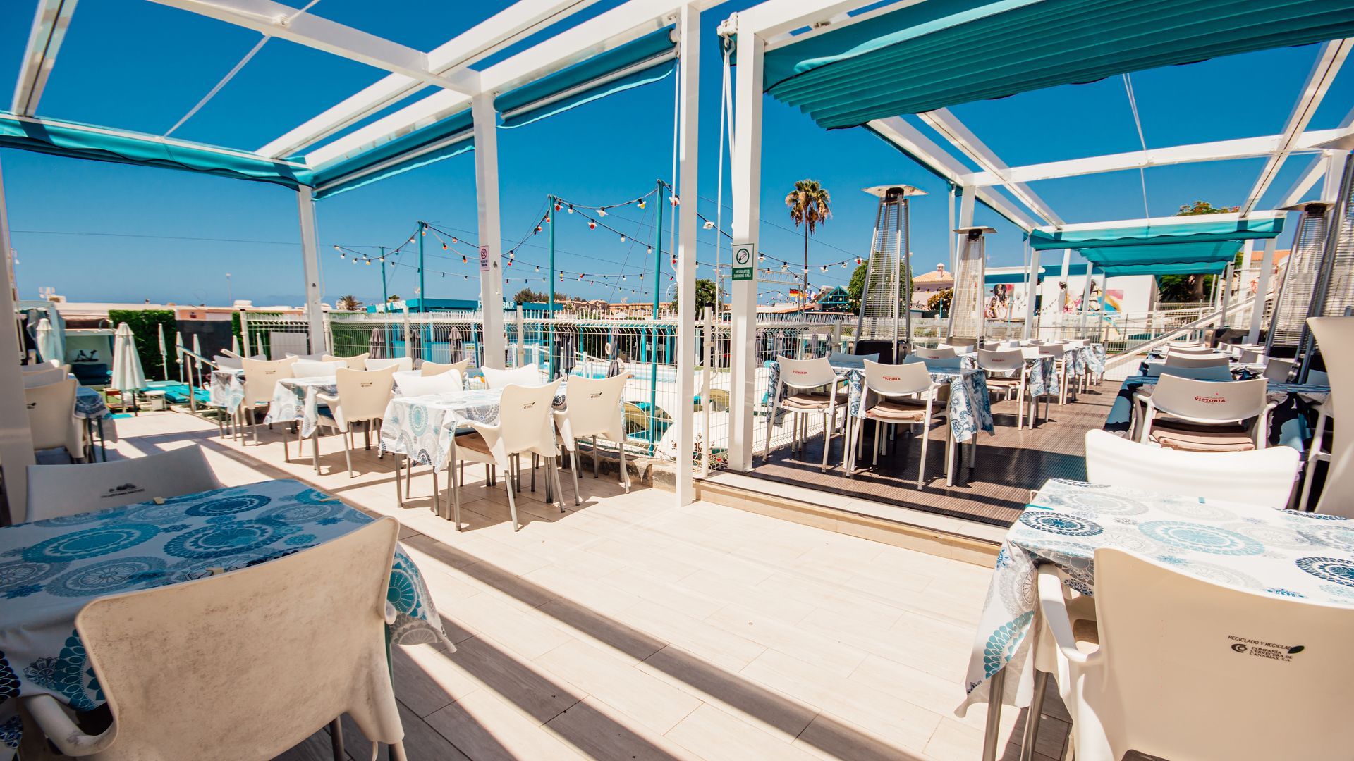 Sunny outdoor seaside café with white tables and chairs under blue-and-white shade canopies.