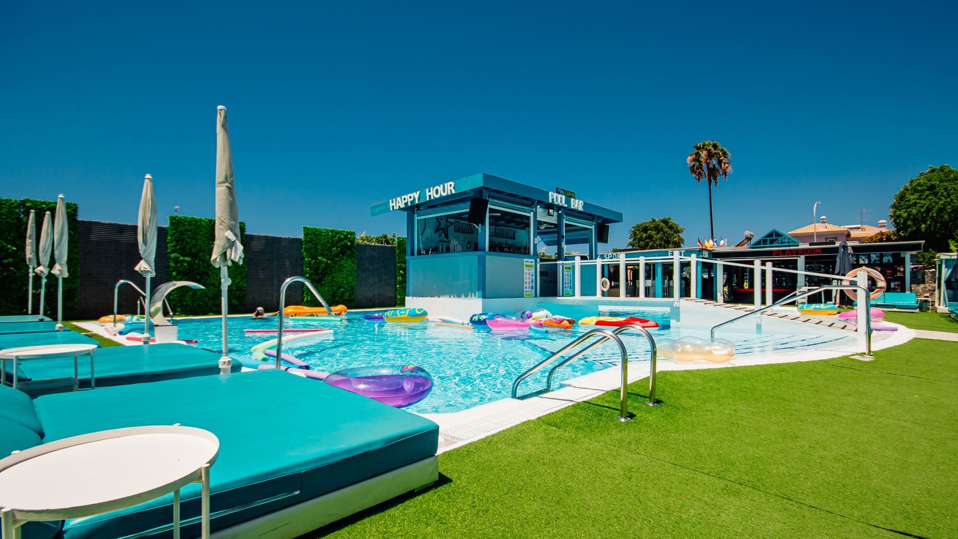 Outdoor swimming pool with turquoise water, umbrellas, and a poolside stage under a clear blue sky