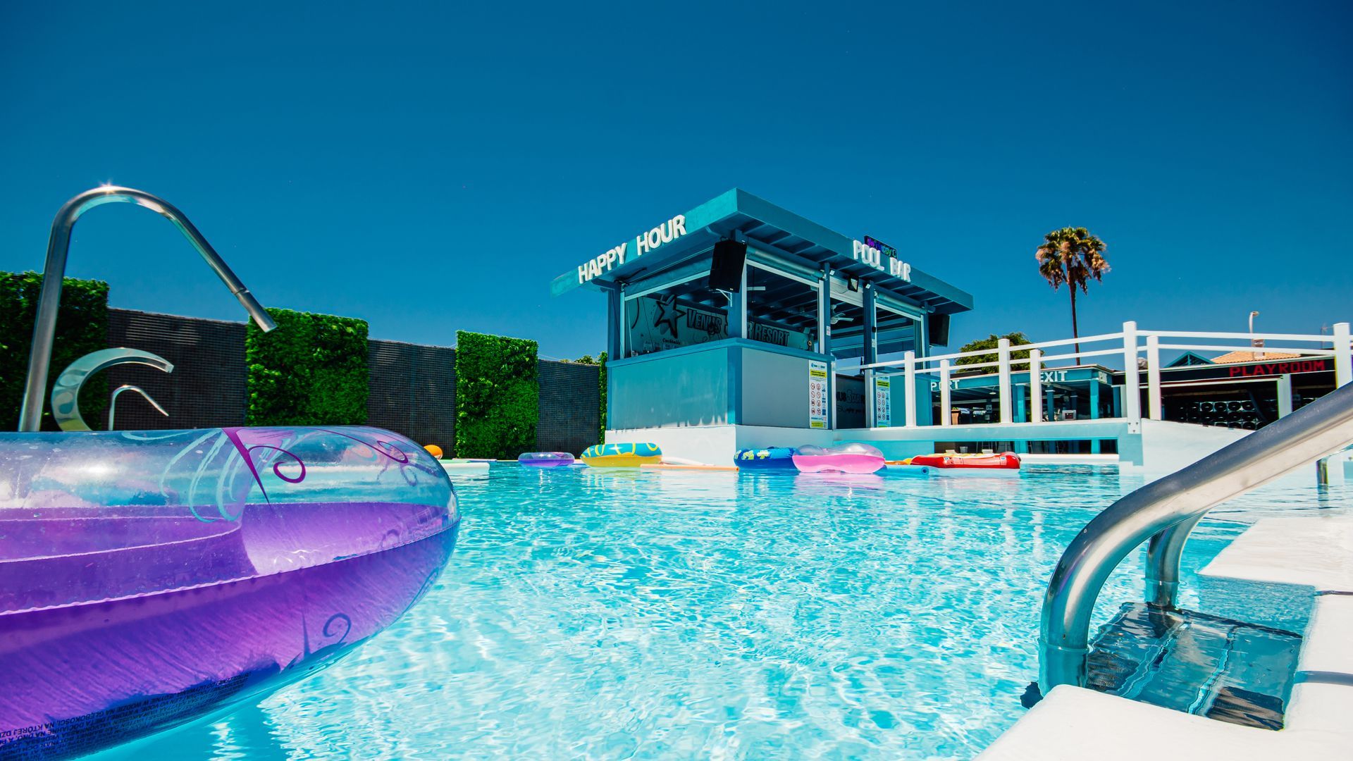 Bright pool with purple float, waterslide, and modern resort building under a clear blue sky