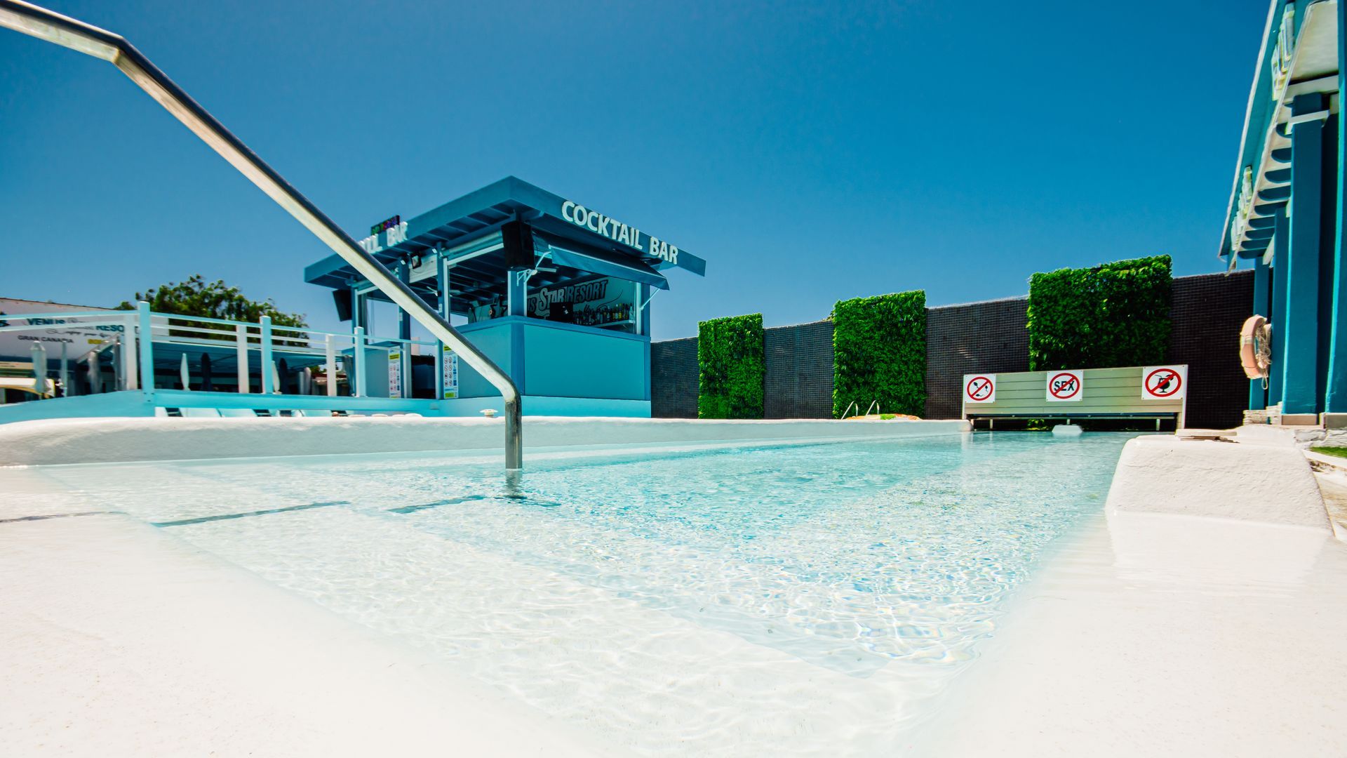 Empty outdoor pool with white water, blue shade structures, and a lifeguard tower under a clear sky