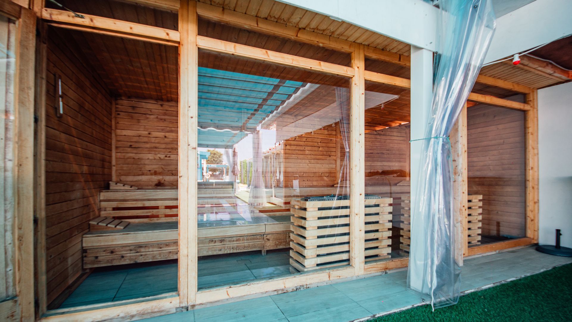 Wooden sauna-style lounge with benches, stacked towels, and hanging curtains by a green floor