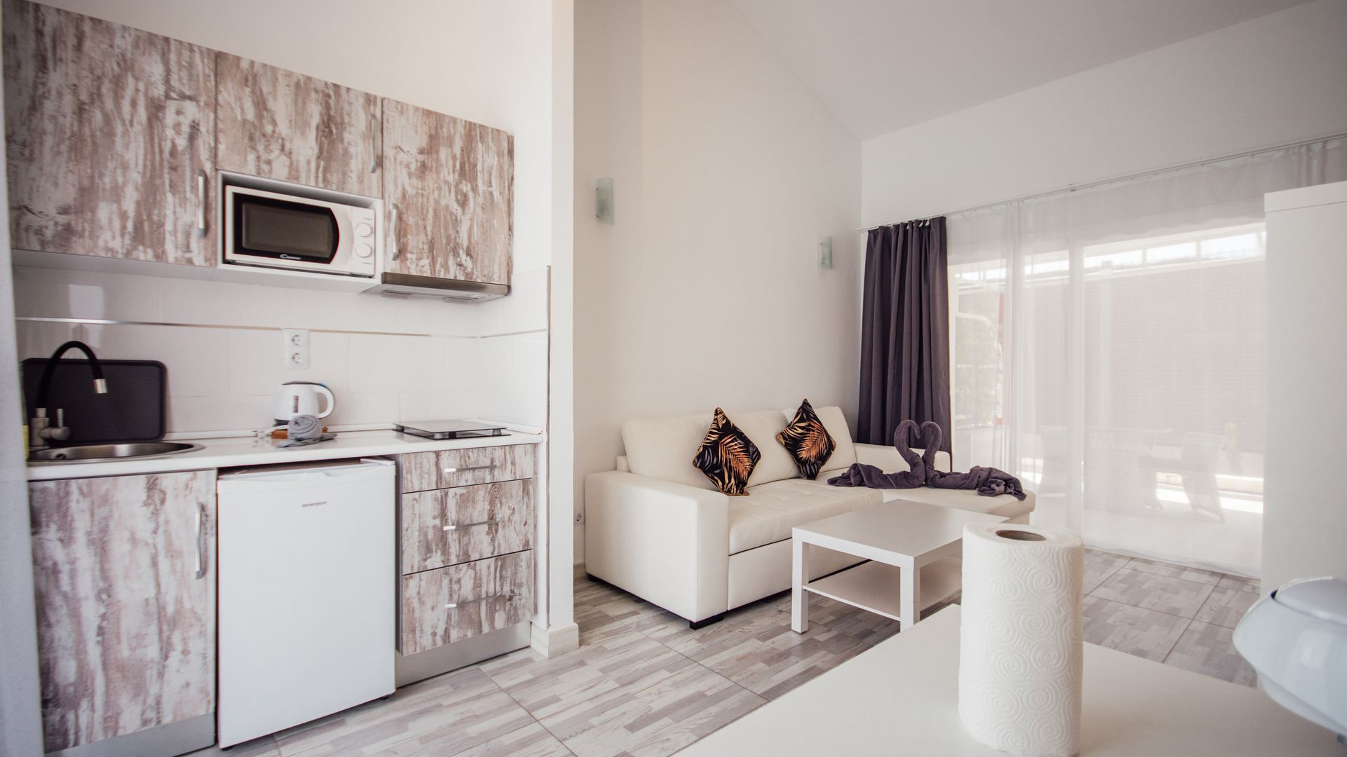 Bright studio apartment with kitchenette, white sofa, coffee table, and large sunlit windows