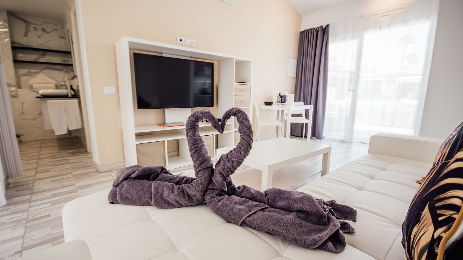 Bright living room with white sectional sofa, TV, and a purple towel folded into a heart shape on the couch