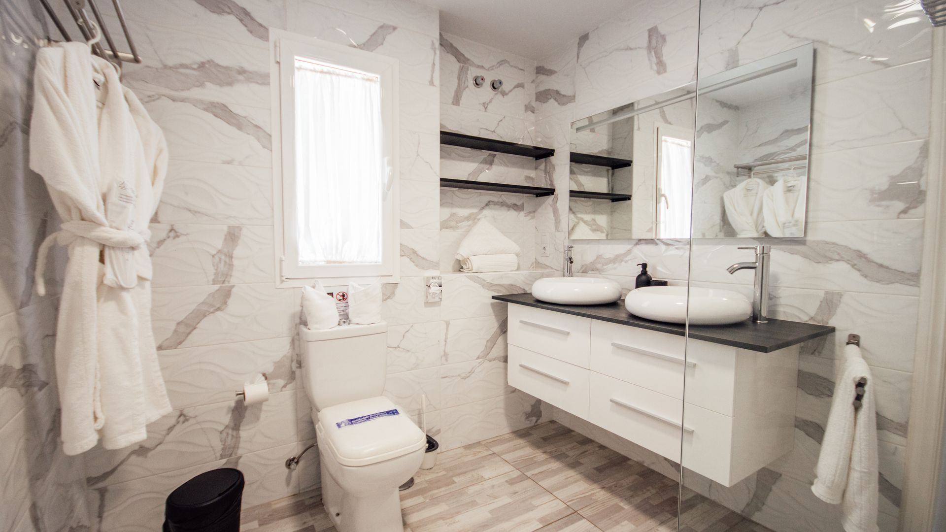 Bright modern bathroom with marble walls, white vanity, round mirrors, and robes hanging on the wall
