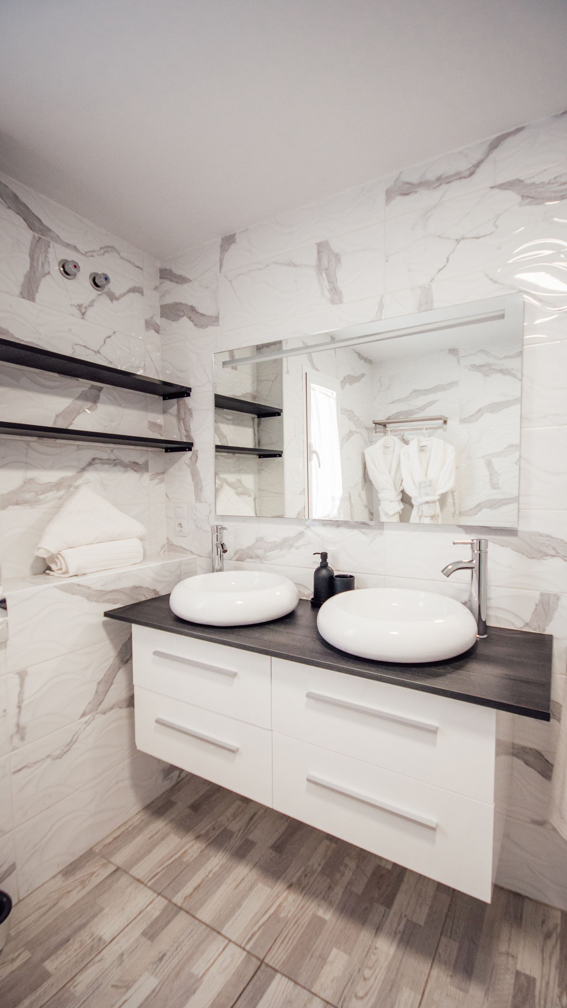 Modern bathroom with double vessel sinks, large mirror, white marble walls, and wood-look floor