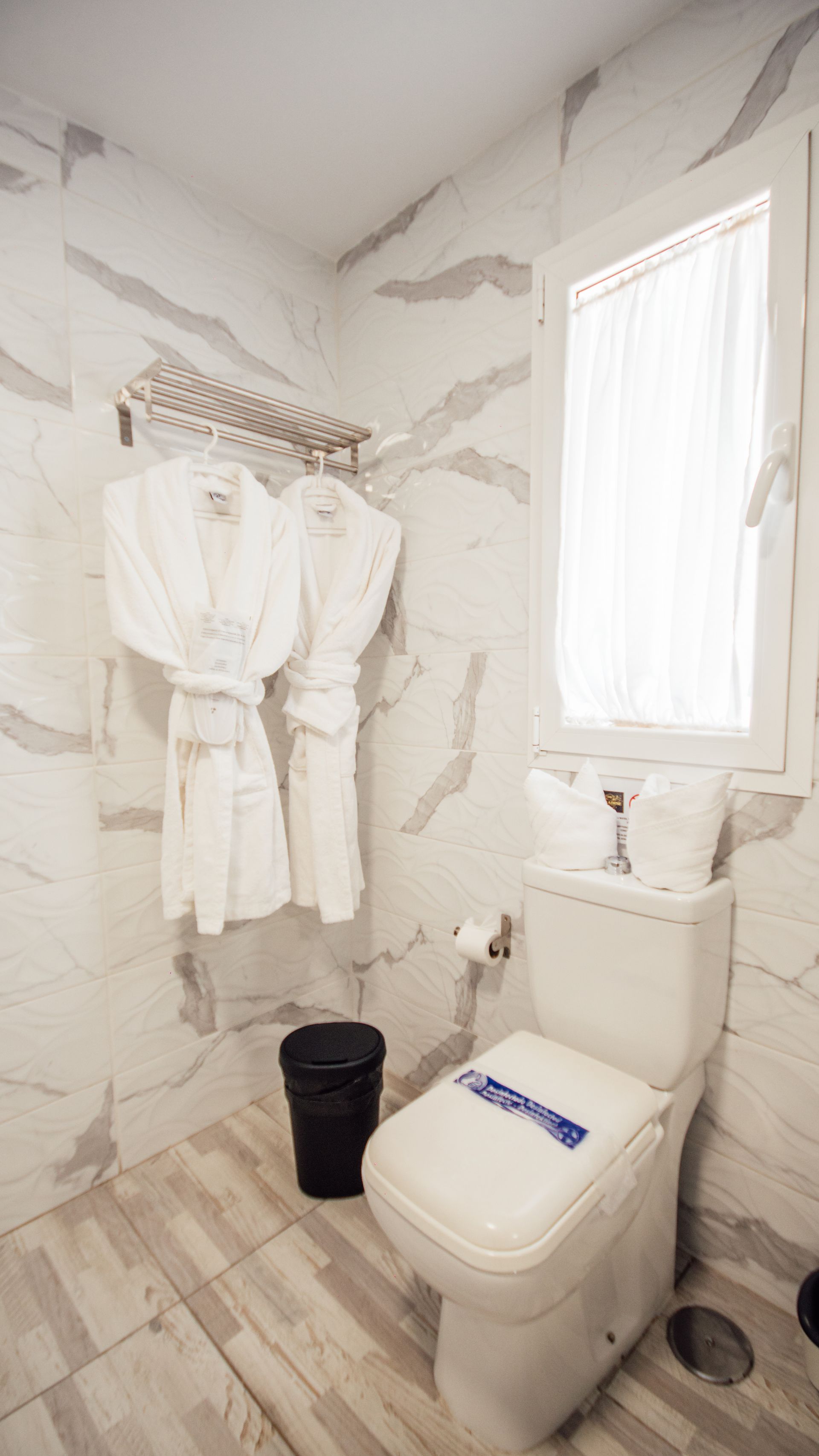 Small white bathroom with marble walls, toilet, window, black bucket, and hanging white robes.