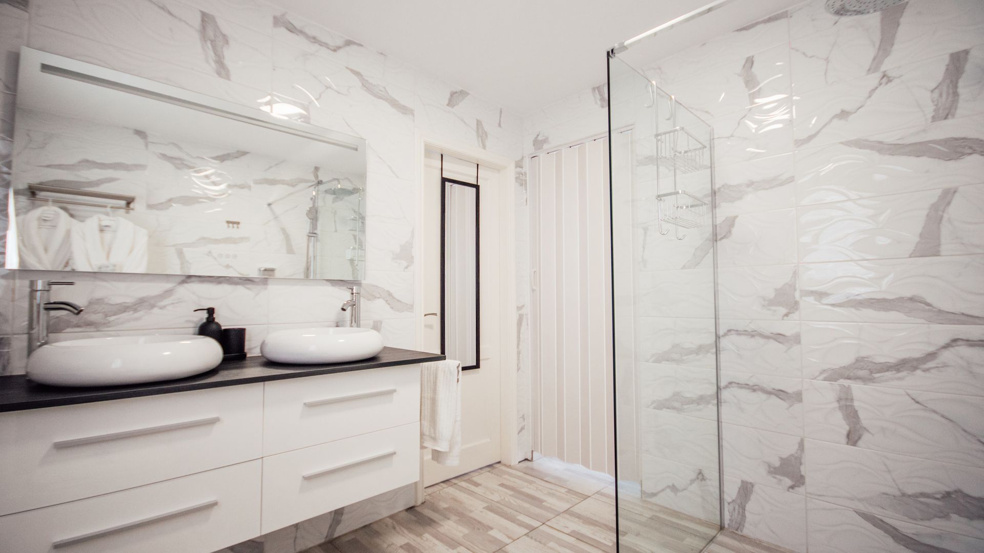 Bright white marble bathroom with double sinks, large mirror, glass shower, and light wood floor.