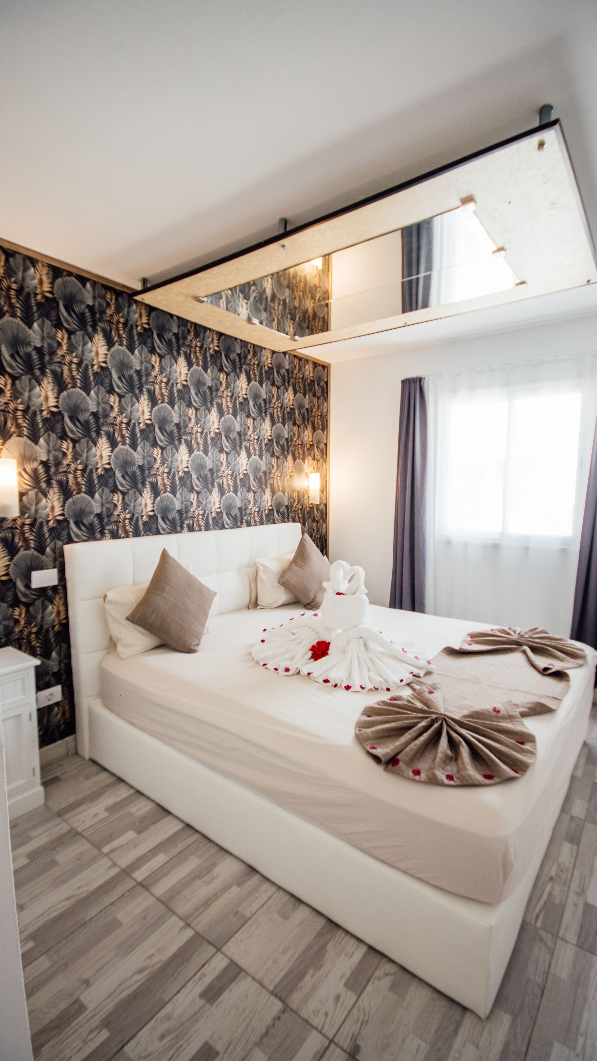 Elegant bedroom with white bed, patterned wallpaper, warm lighting, and large mirror ceiling detail