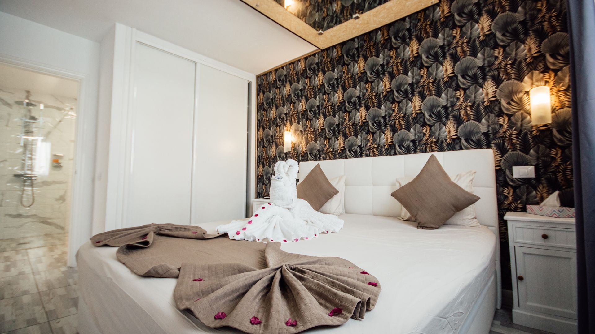 A neatly made bed with white bedding, taupe pillows, folded towels, and dark patterned wallpaper in a cozy bedroom.