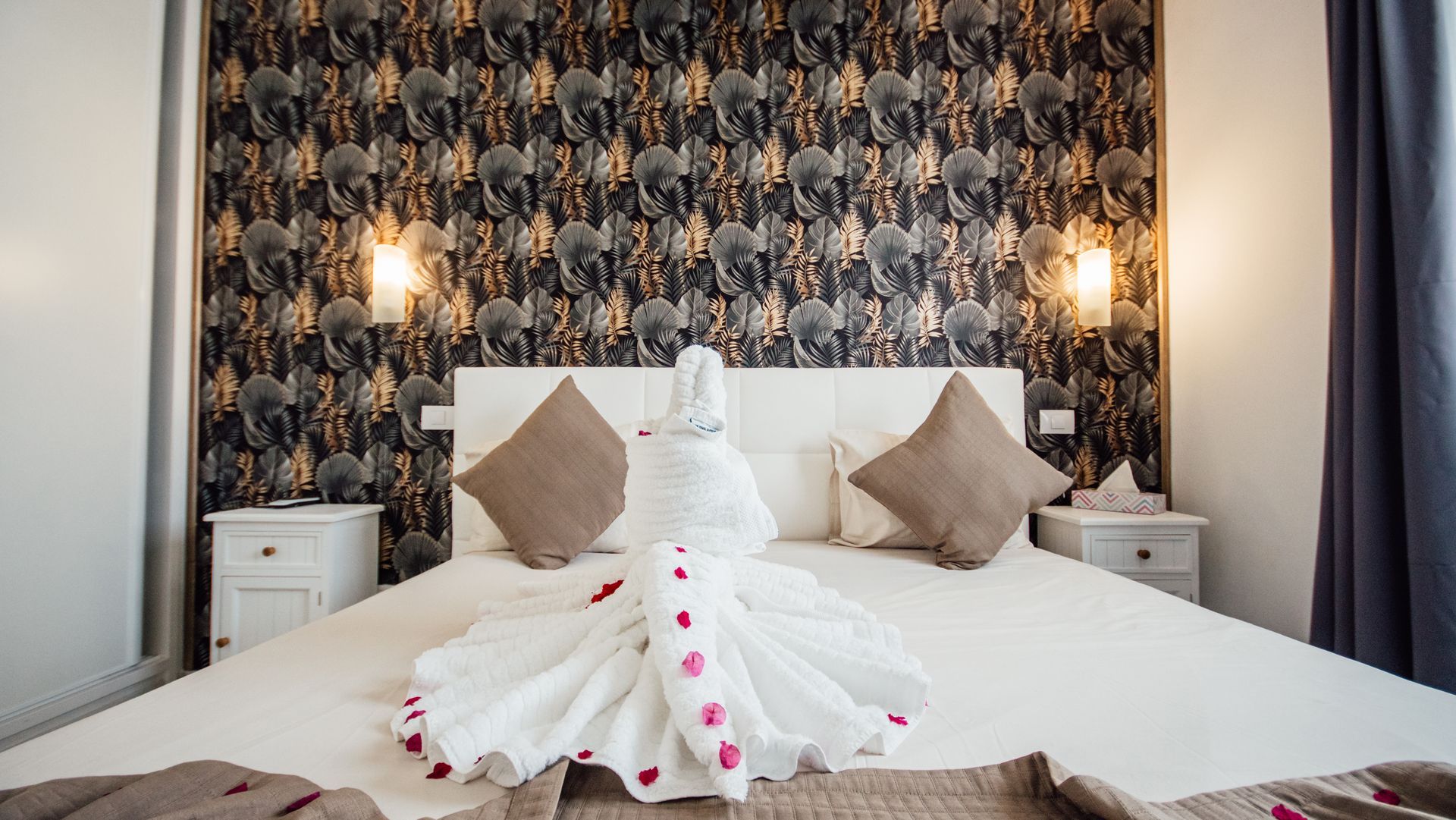 White wedding dress on a bed in a patterned hotel room with rose petals and bedside lamps