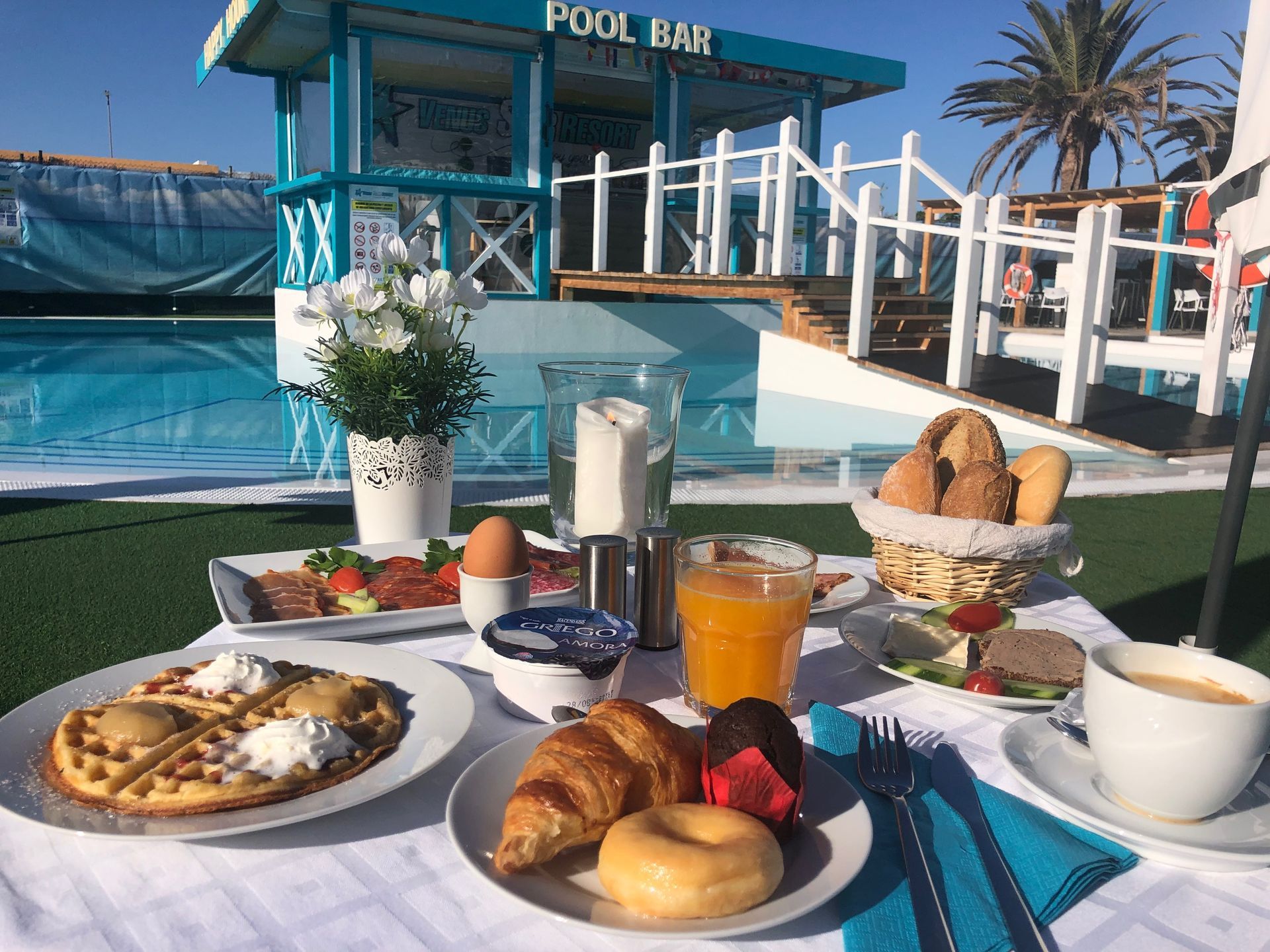 Breakfast spread on a poolside table with pastries, fruit, coffee, and juice at a pool bar.