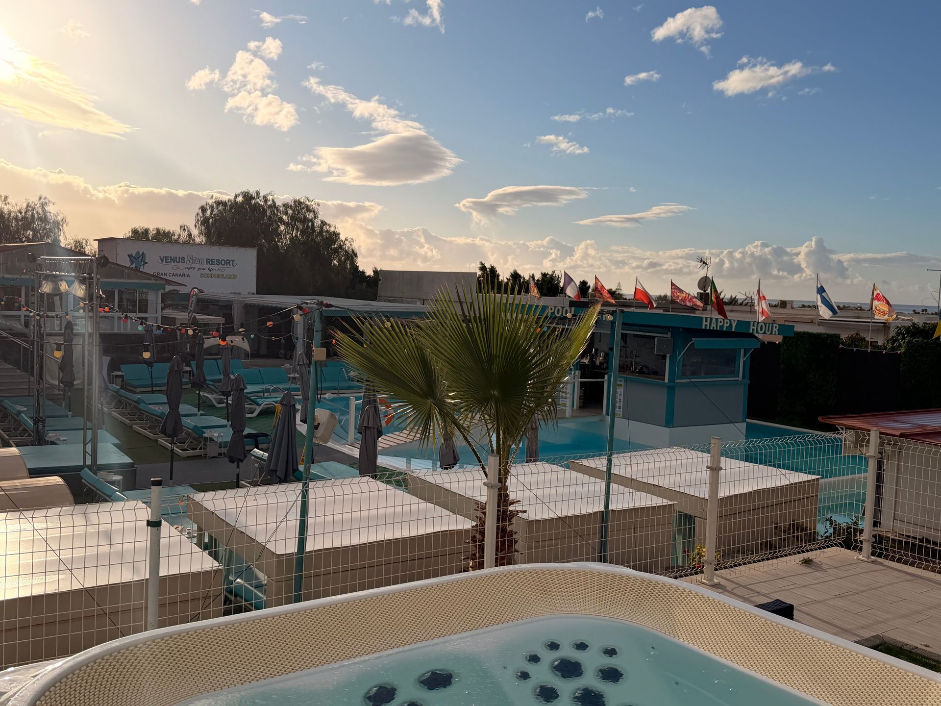 Sunny resort pool with lounge chairs, palm trees, and flags along a seaside deck at sunset