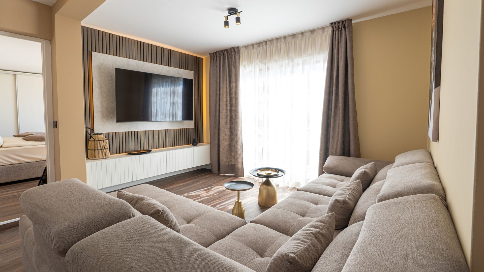 Modern living room with a large sectional sofa, wall-mounted TV, and floor-to-ceiling curtains by a bright window