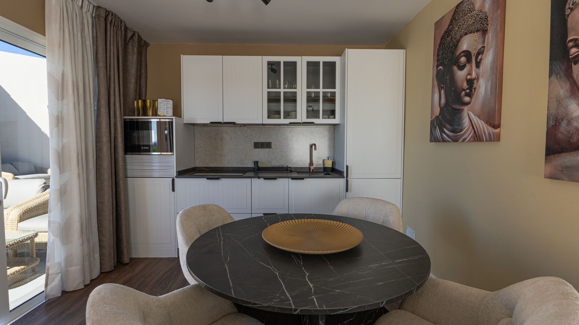 Modern dining area with round black table, beige chairs, and compact kitchen by a curtained window.