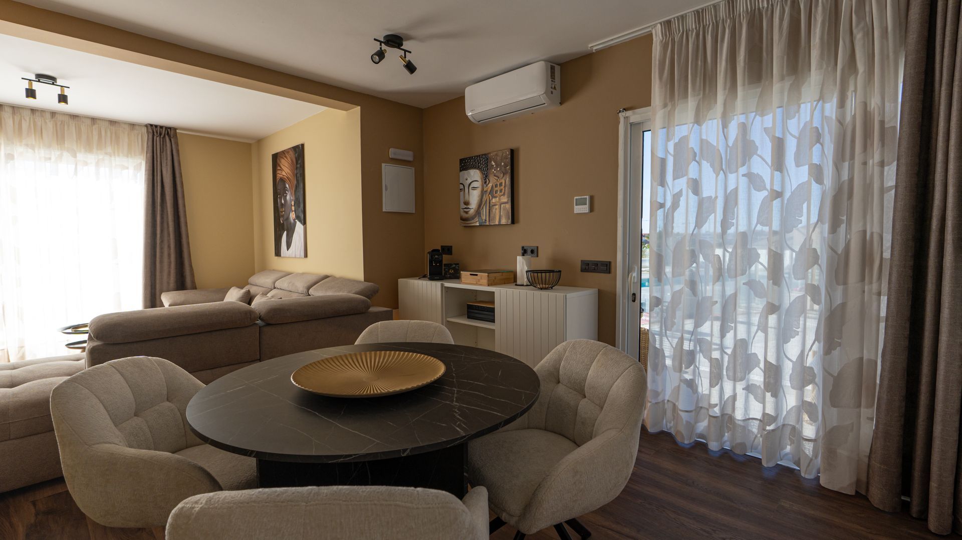 Bright living-dining room with round table, beige chairs, sofa, and sunlight through sheer curtains