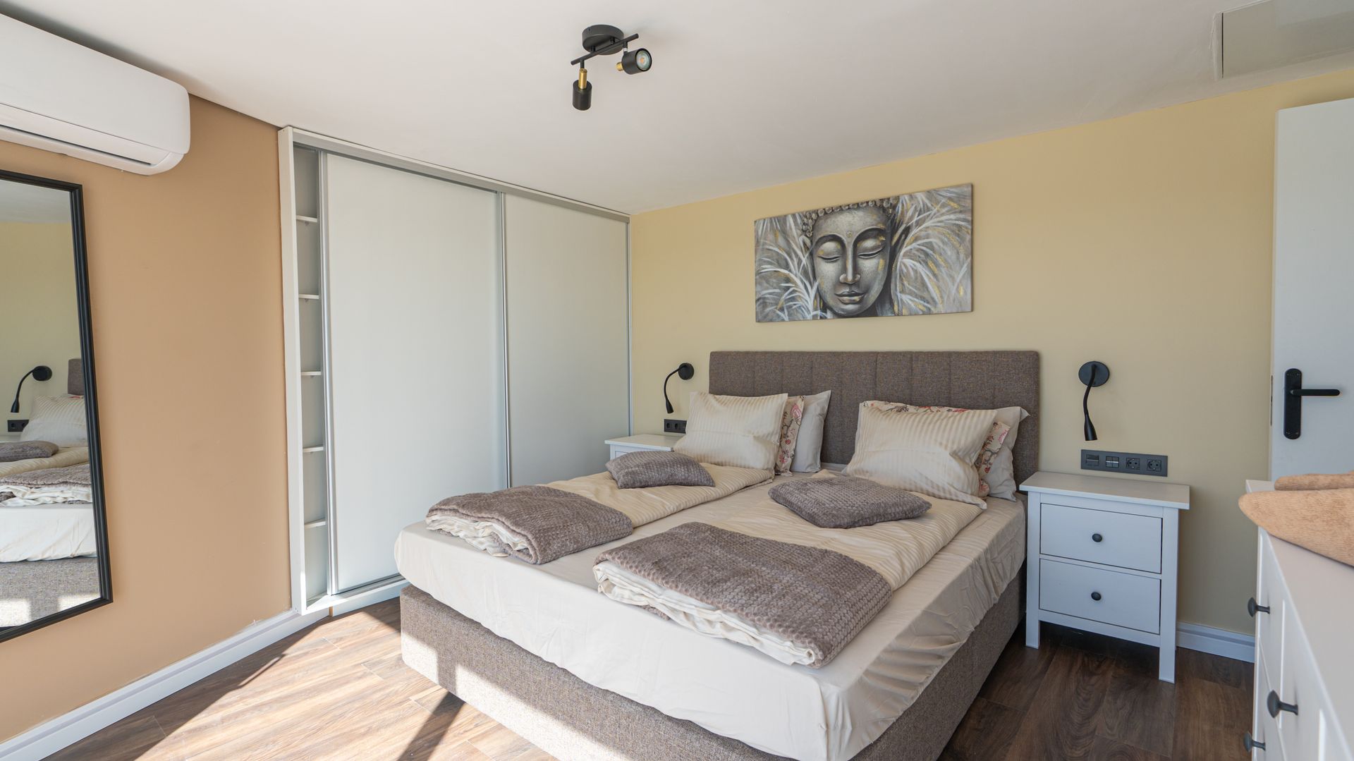 Bright bedroom with a bed, mirrored wardrobe, nightstand, wall art, and sunlight through a window.
