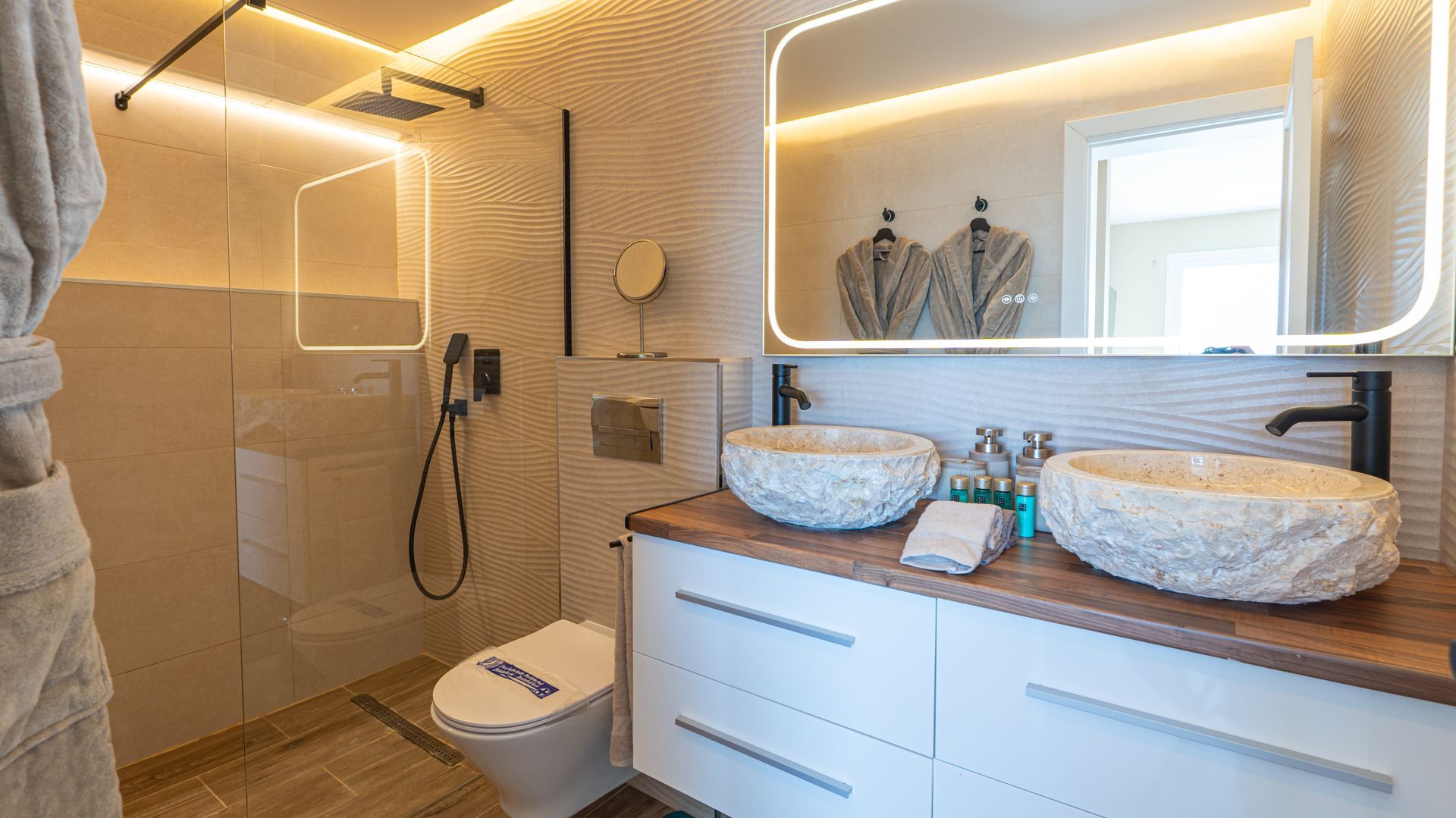 Modern bathroom with glass shower, illuminated mirror, double sink vanity, and warm beige tile walls