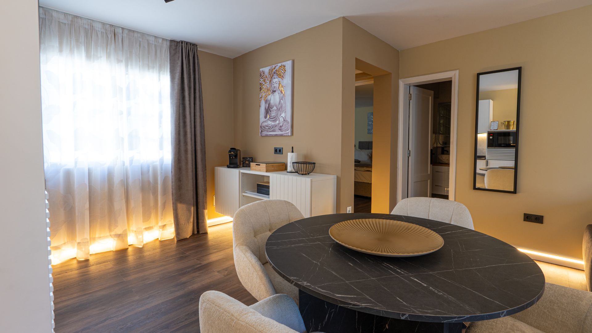 Modern dining room with round table, beige chairs, mirror, and sheer curtains glowing with warm light