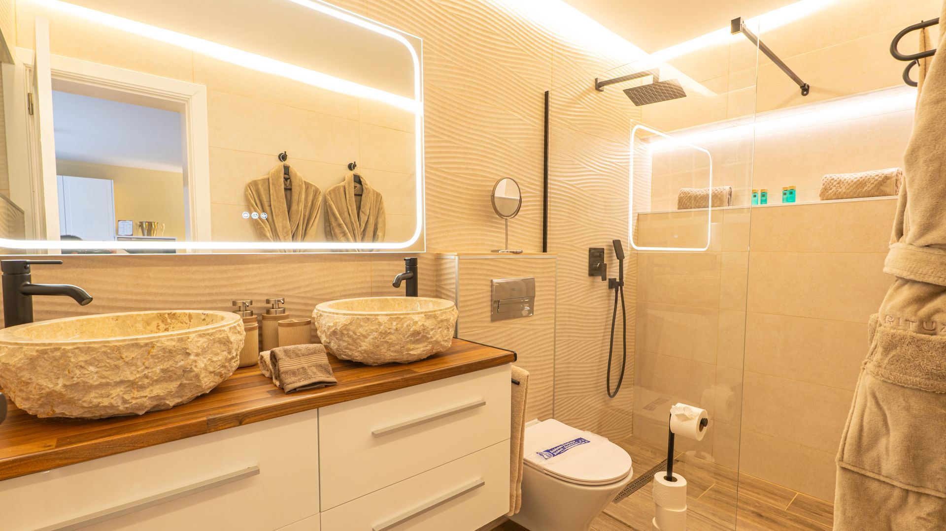Warm beige bathroom with illuminated mirror, marble sink counter, glass shower, and towels hanging on the right.