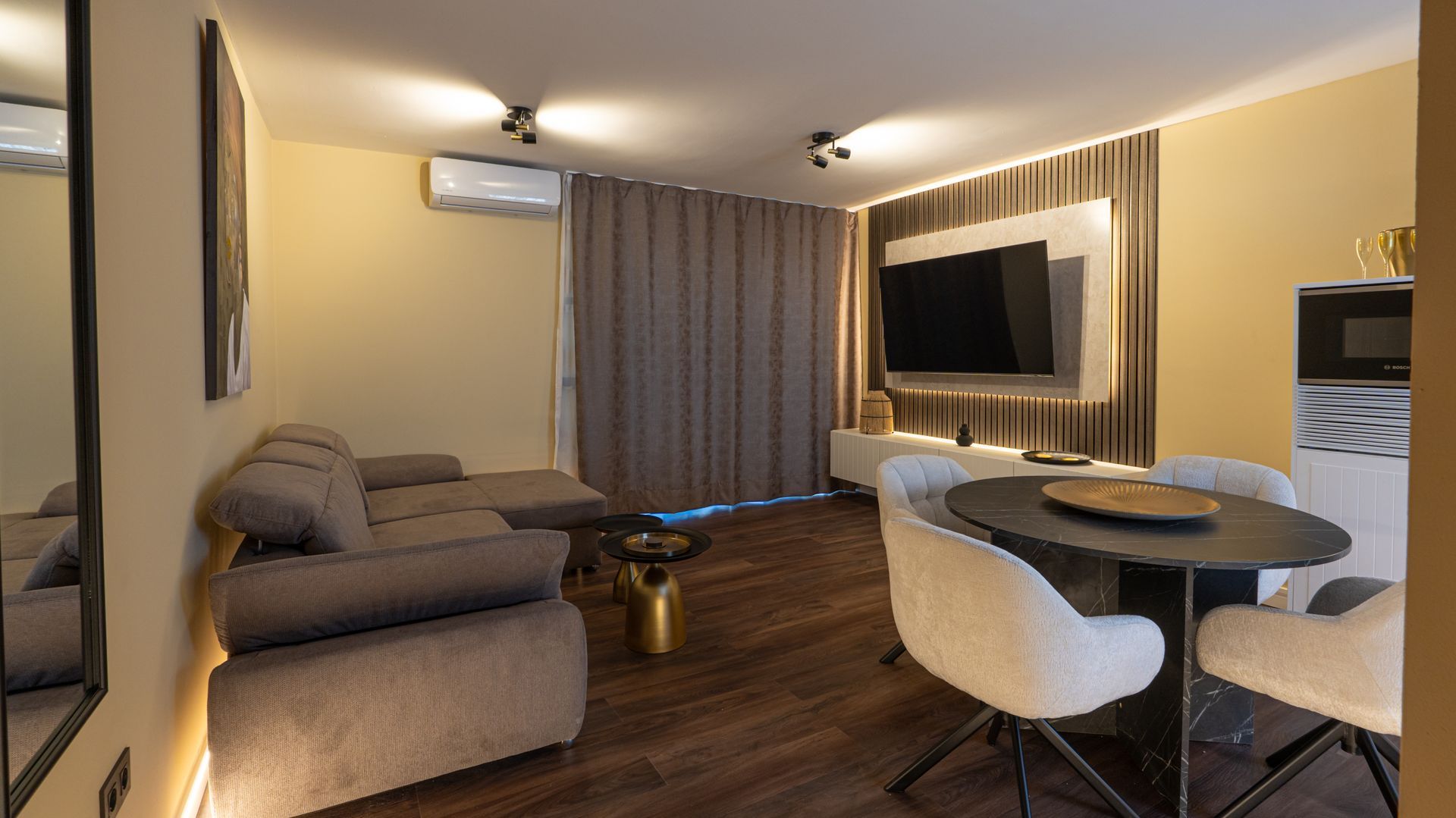 Modern living room with gray sofa, round dining table, wall-mounted TV, and curtains.