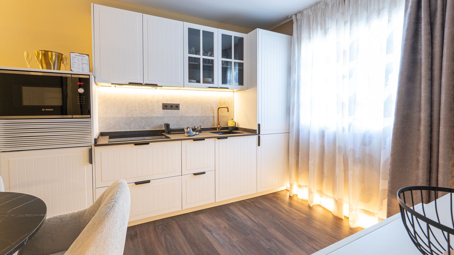 Bright modern kitchenette with white cabinets, stove, sink, and sunlight through sheer curtains