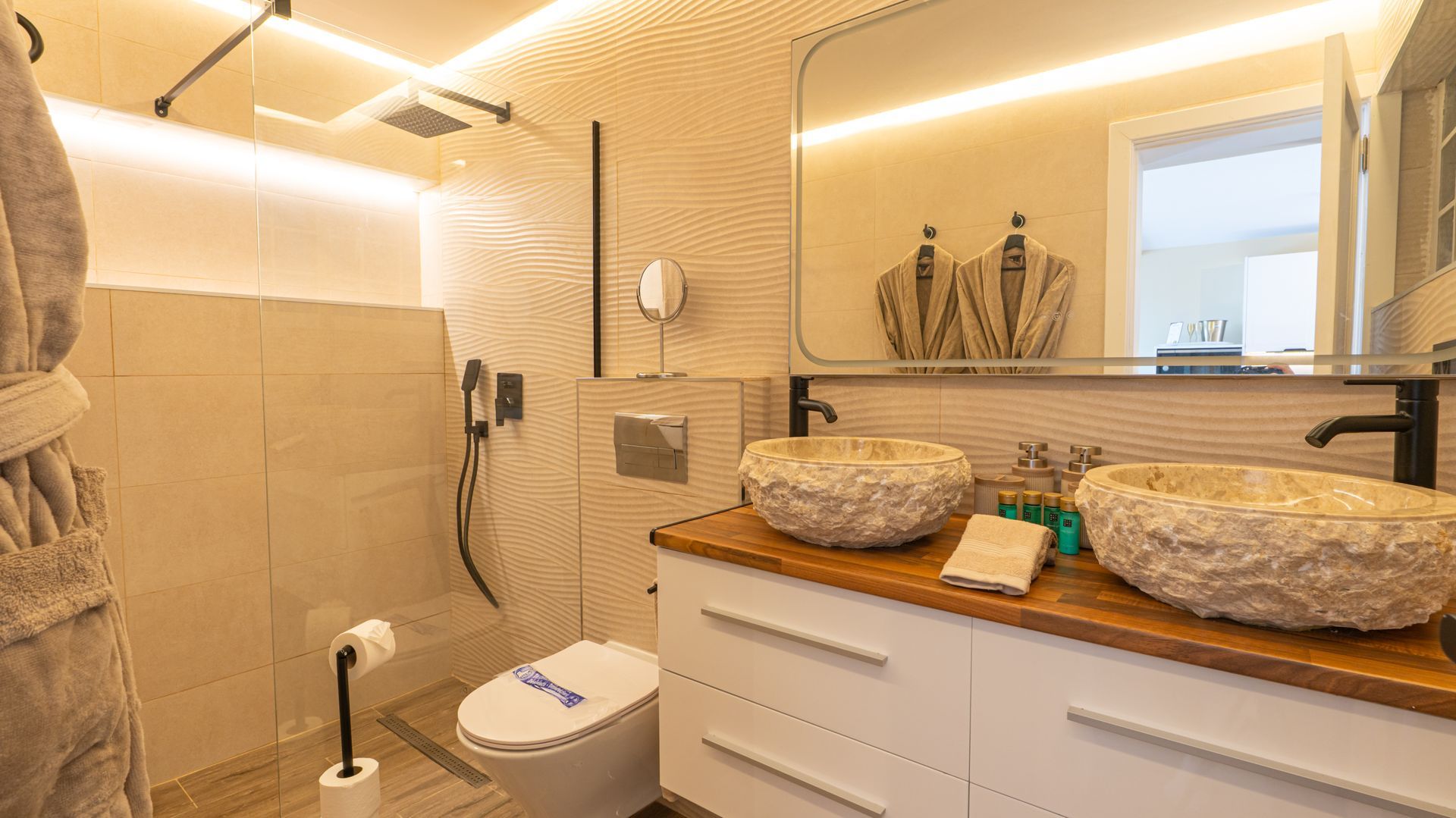 Modern beige bathroom with twin stone sinks, large mirror, and a toilet beside a shower area.