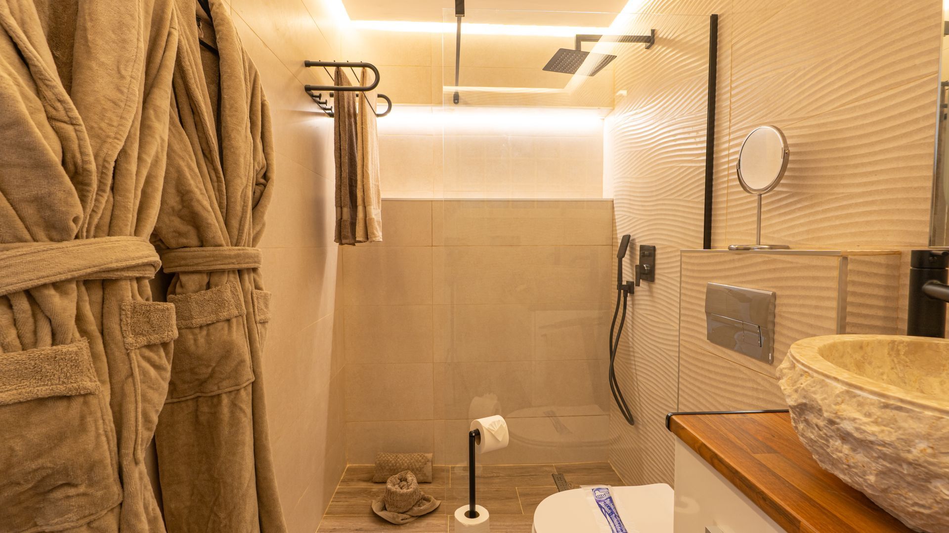 Warm beige tiled shower with handheld showerhead, wall shelf, and towels in a modern bathroom.
