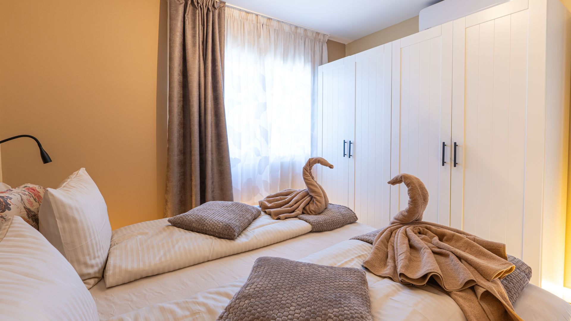 Bright bedroom with white wardrobes, two beds, curtains, and towel swans on the bed