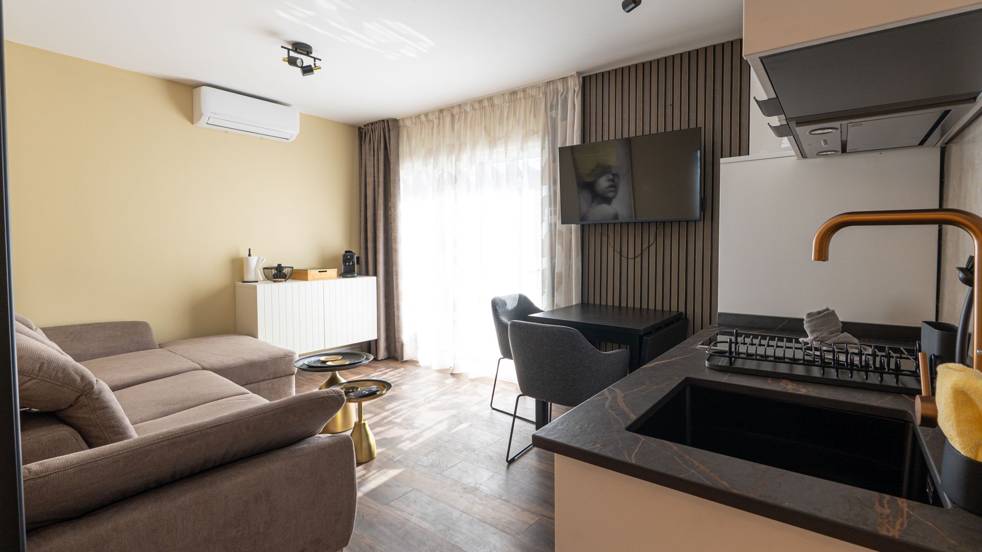 Modern living room with beige sofa, wall-mounted TV, dining table, and open kitchen.