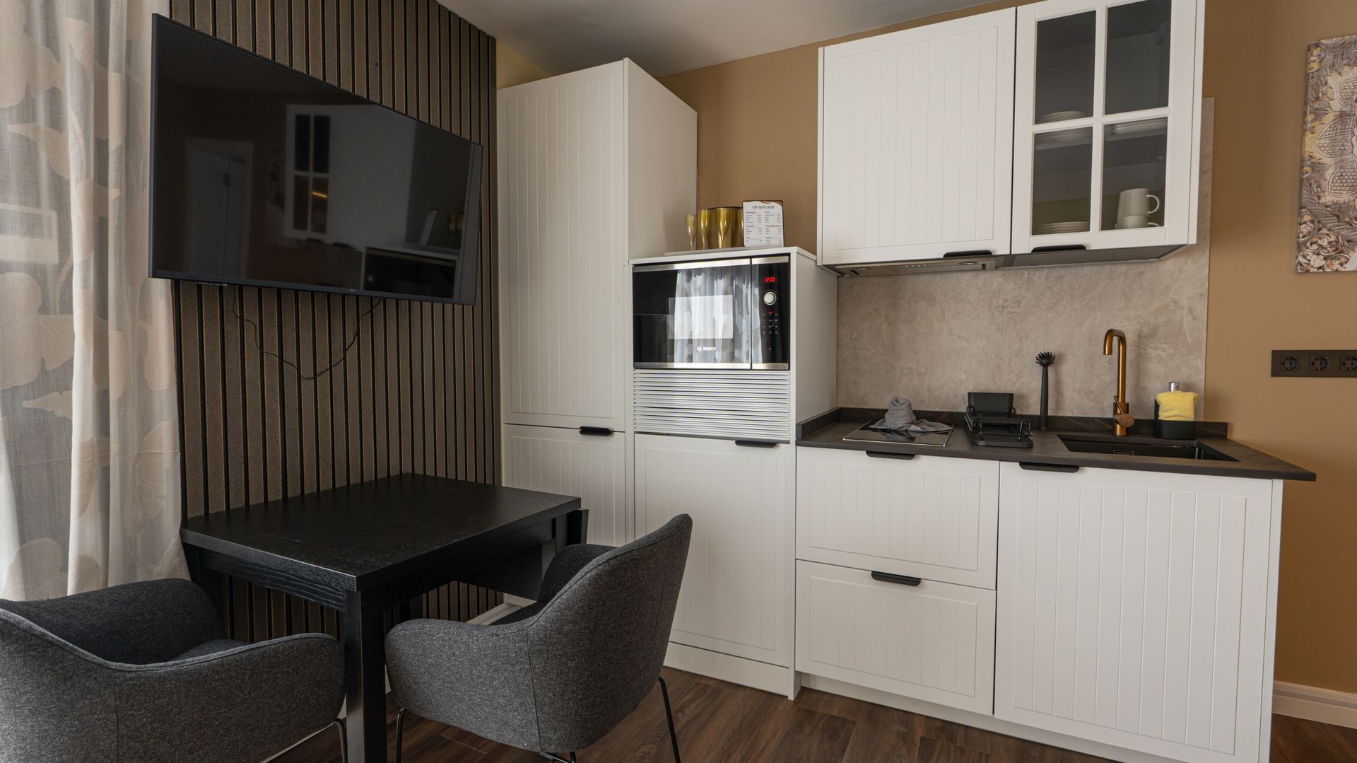 Compact modern kitchenette and dining nook with white cabinets, black table, and wall-mounted TV