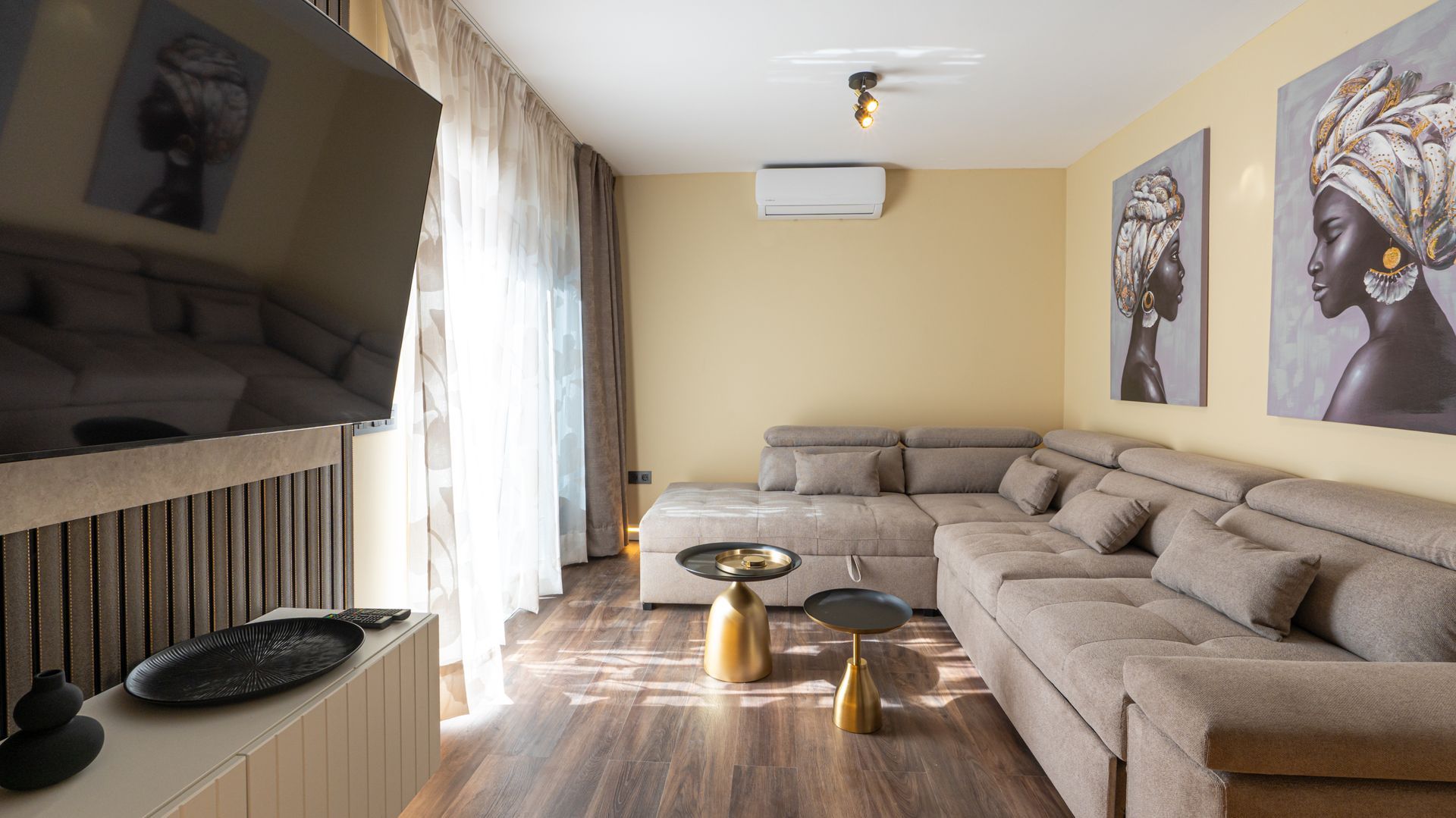 Modern beige living room with sectional sofa, wall-mounted TV, coffee table, and large window with curtains