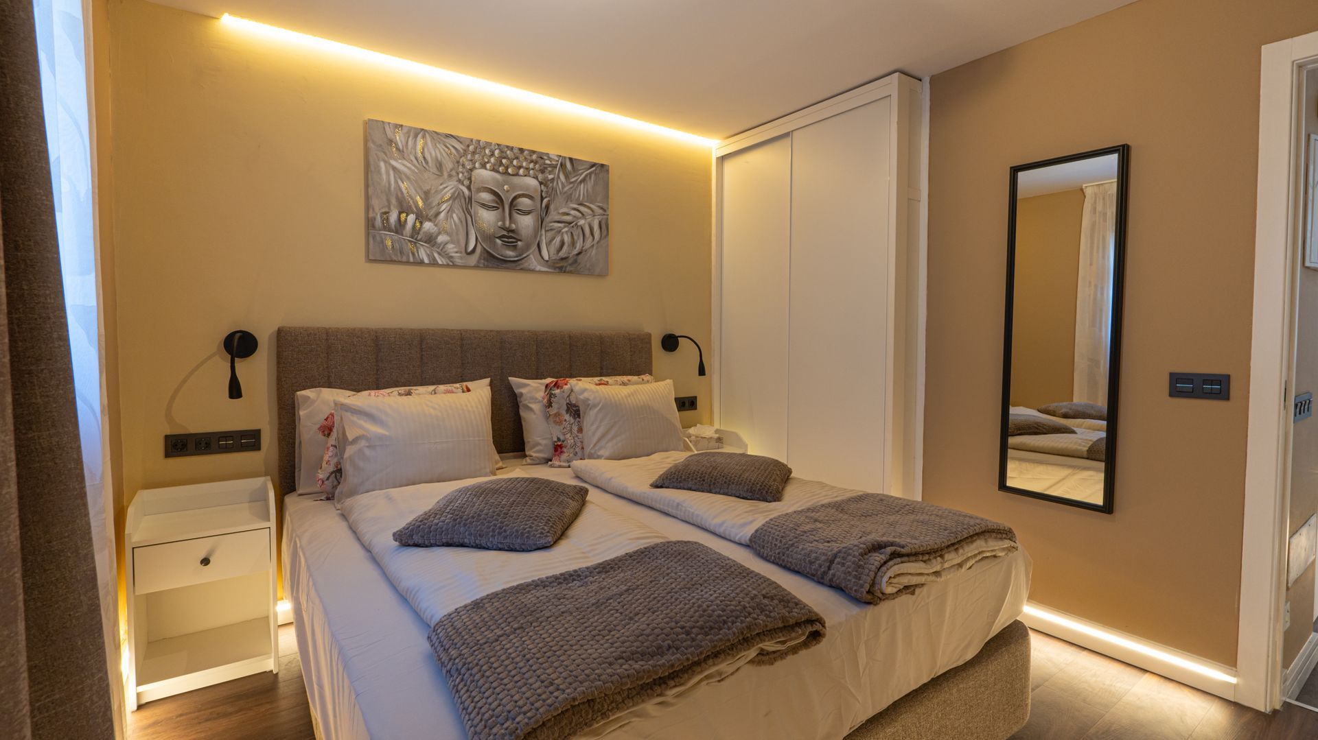 Modern bedroom with a neatly made bed, warm LED lighting, nightstand, wall art, and mirror.