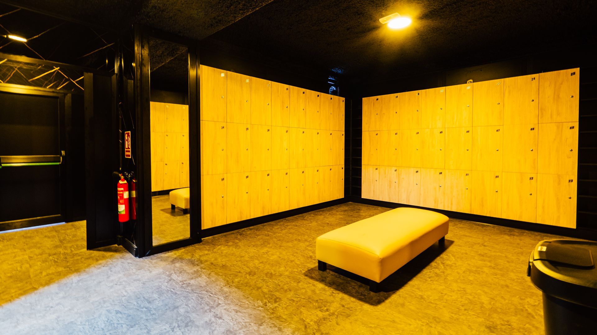 Empty lounge with yellow wall panels, black ceiling, and a central beige bench under warm lighting