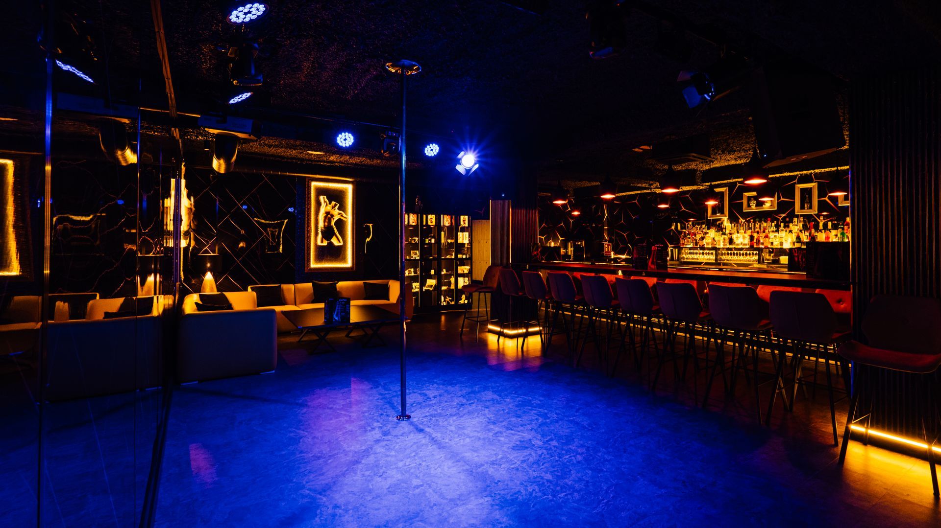 Dimly lit nightclub interior with blue and amber lights, empty dance floor, and lounge seating.