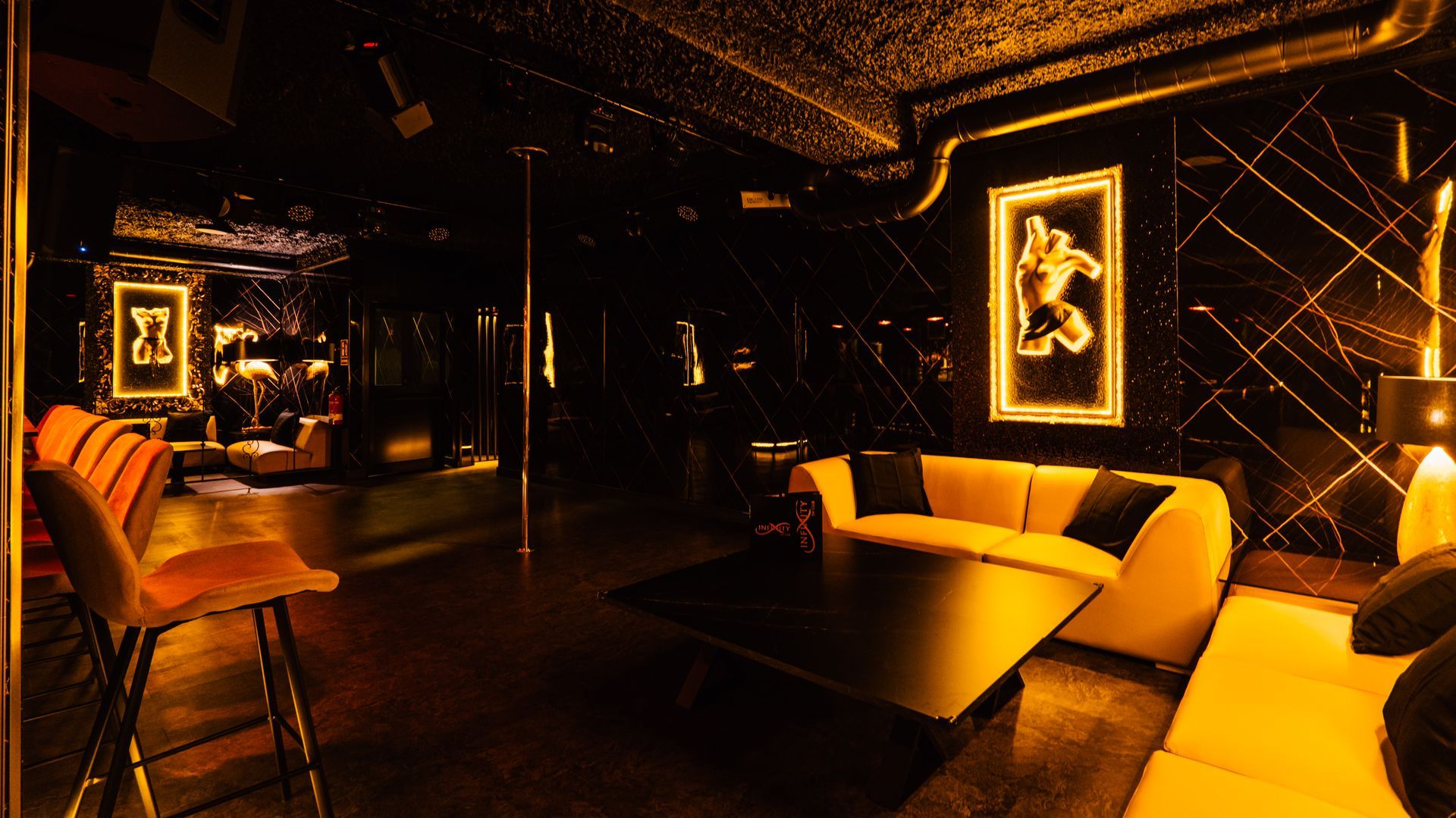 Dim lounge with orange chairs, tables, and wall art lit by warm amber lighting