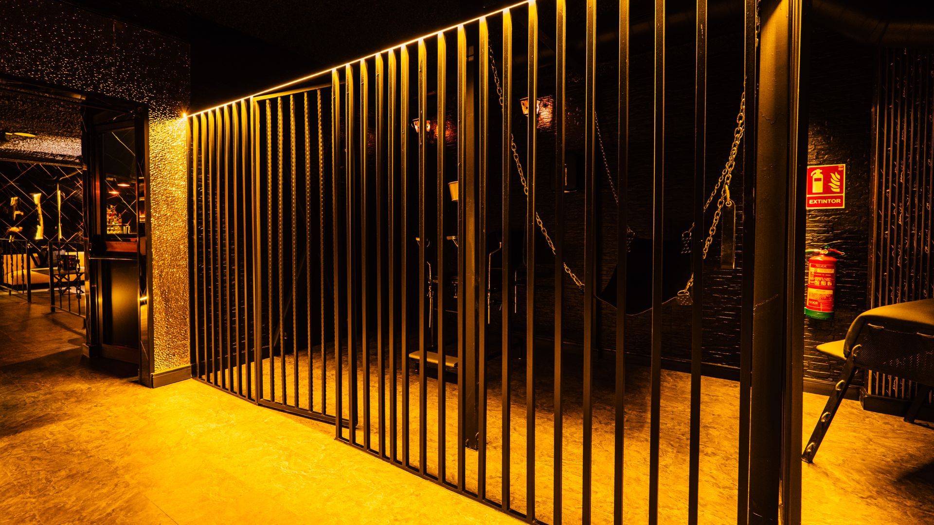 Golden-lit metal gate along a dim walkway at night, with warm orange lighting and dark shadows.
