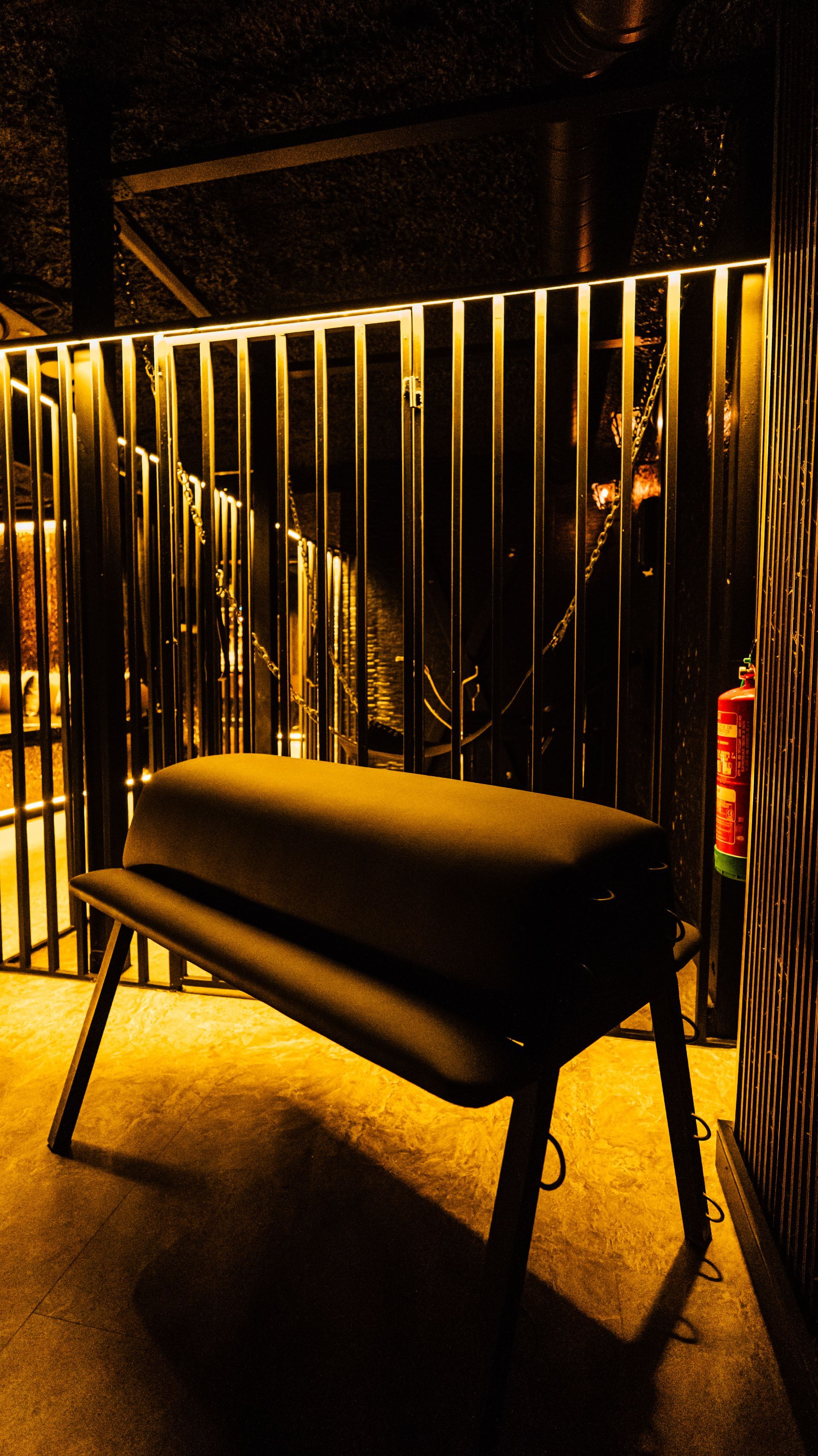 Black bench in front of vertical metal bars, lit by warm amber lighting in a dark setting