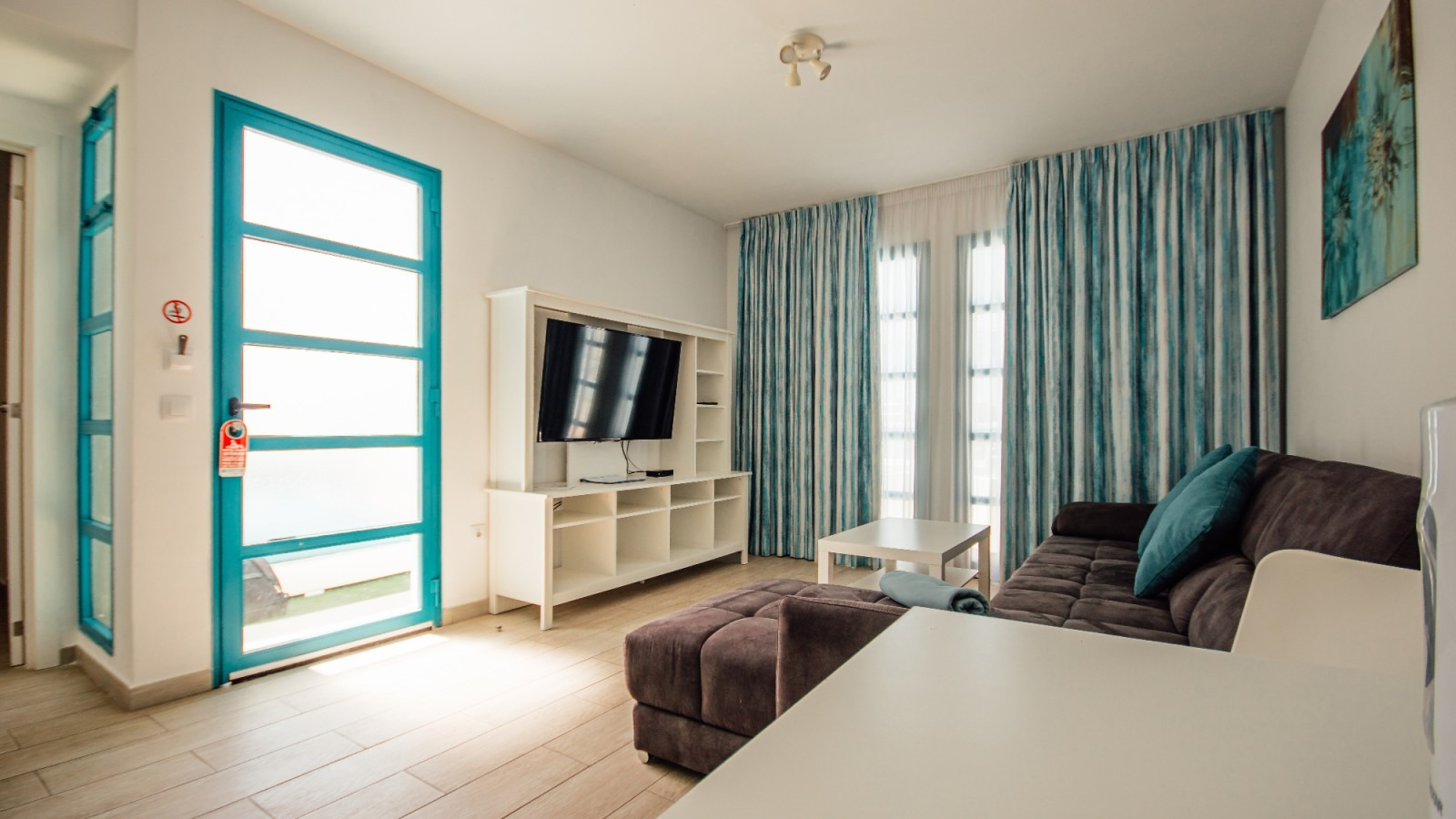 Bright living room with teal door, wall-mounted TV, sofa, ottoman, and blue curtains by a window.
