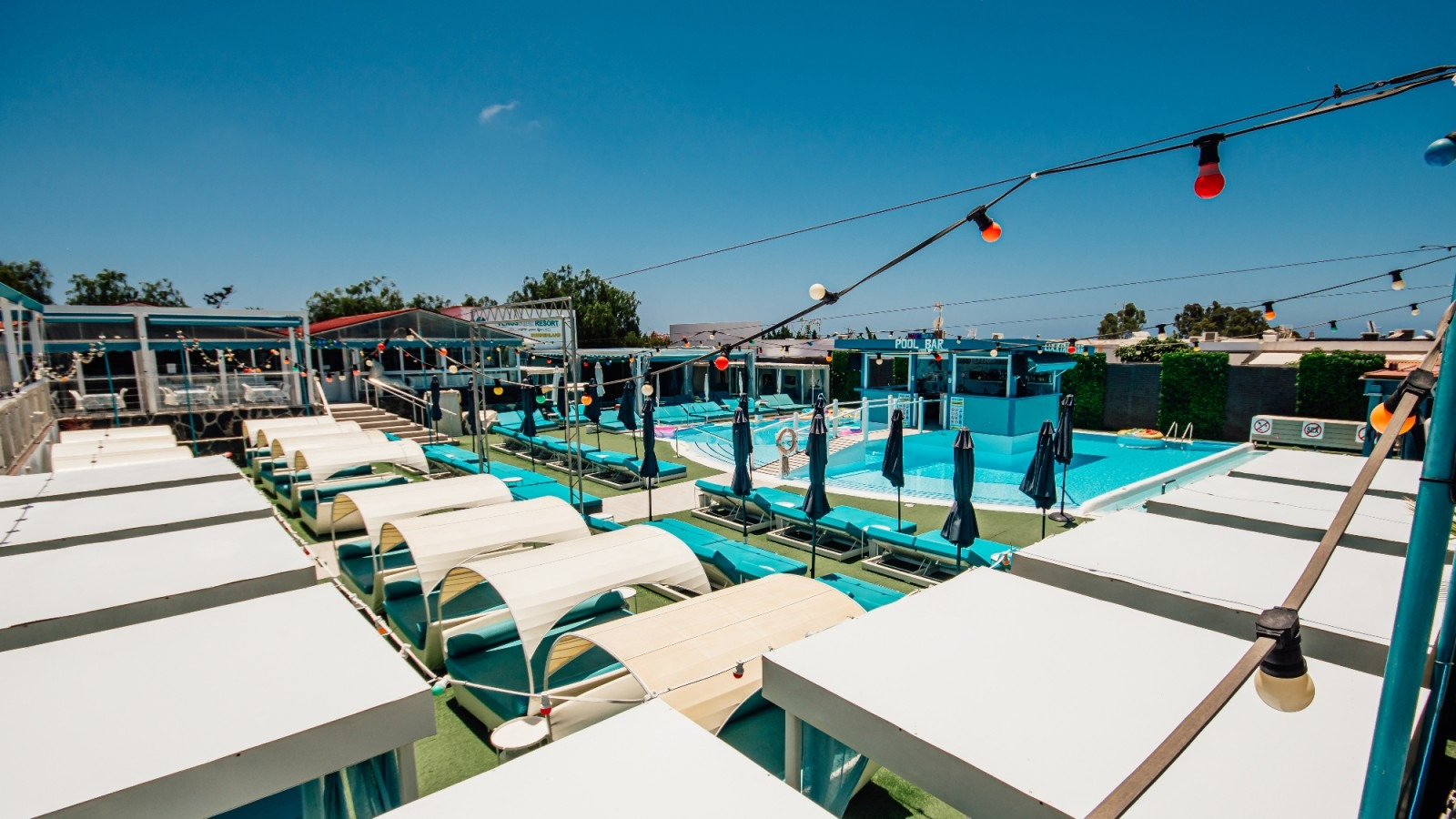 Rooftop pool deck with turquoise water, white lounge chairs, and string lights under a clear blue sky