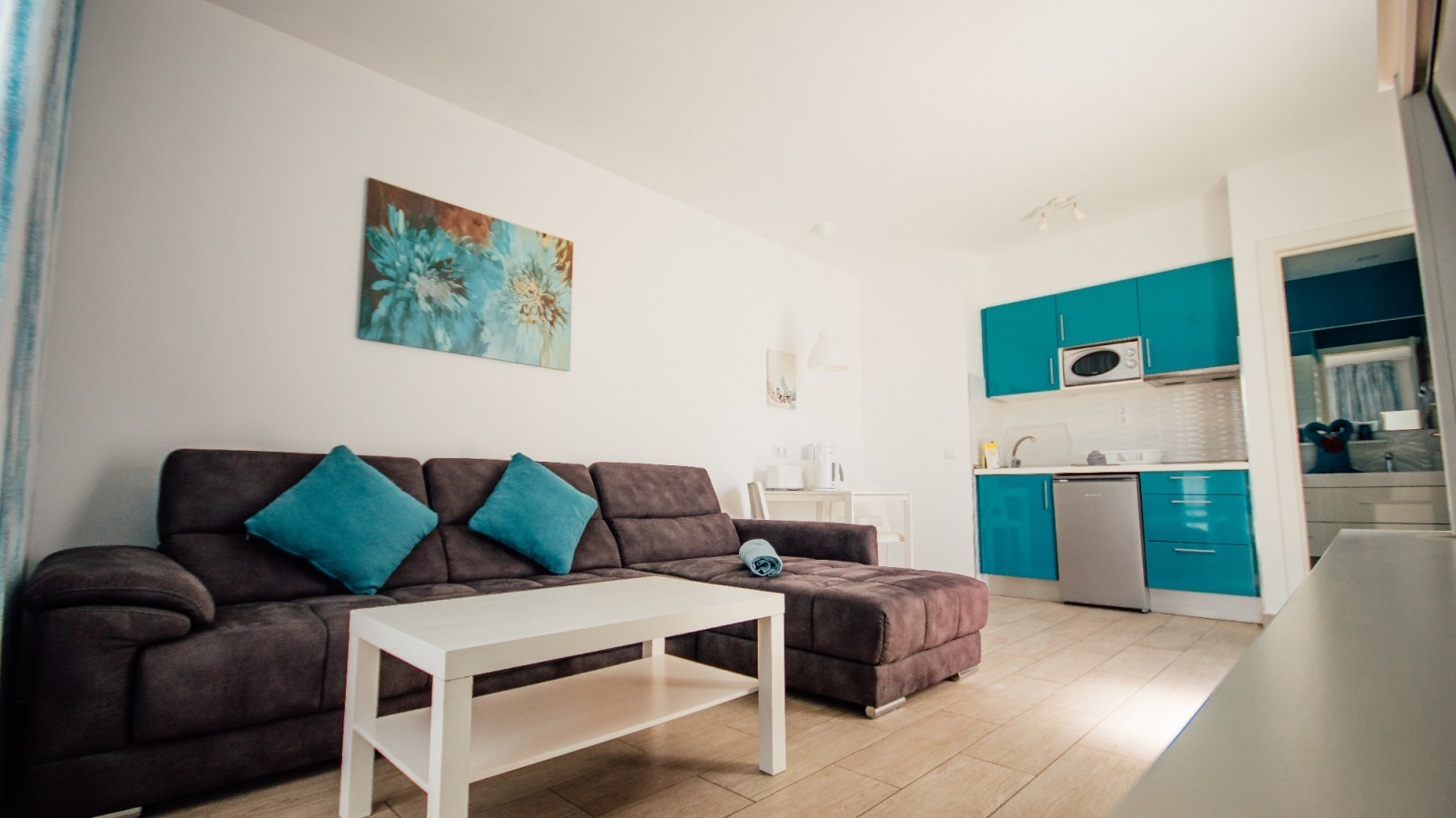 Modern living room with gray sofa, teal cushions, white coffee table, and turquoise kitchen cabinets.