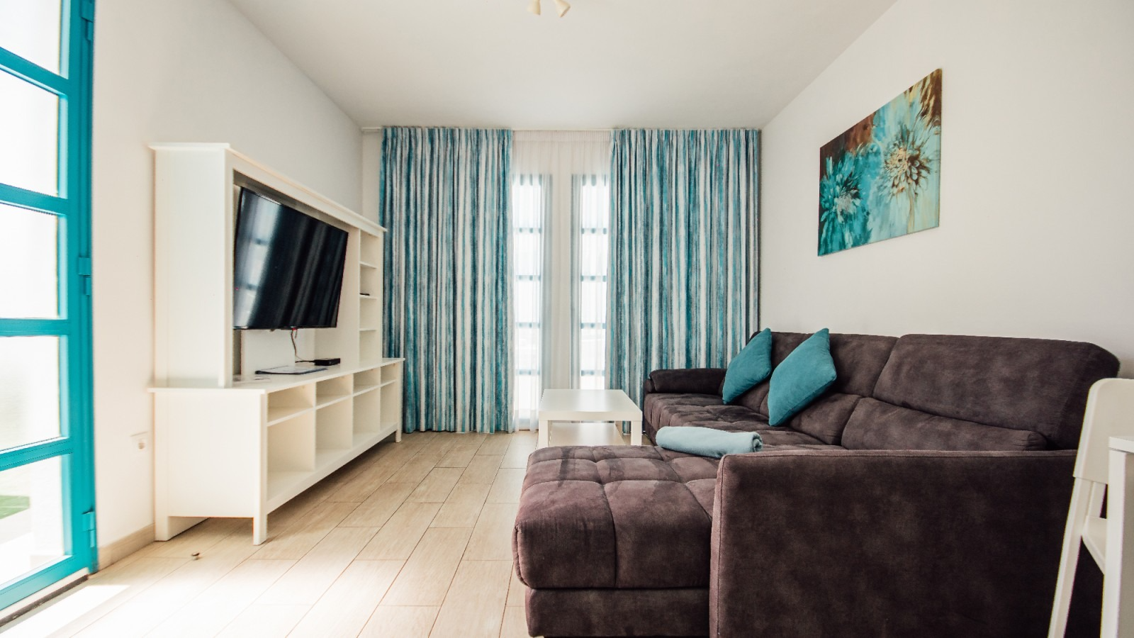 Modern living room with dark sectional, wall-mounted TV, white console, and blue curtains by a bright window