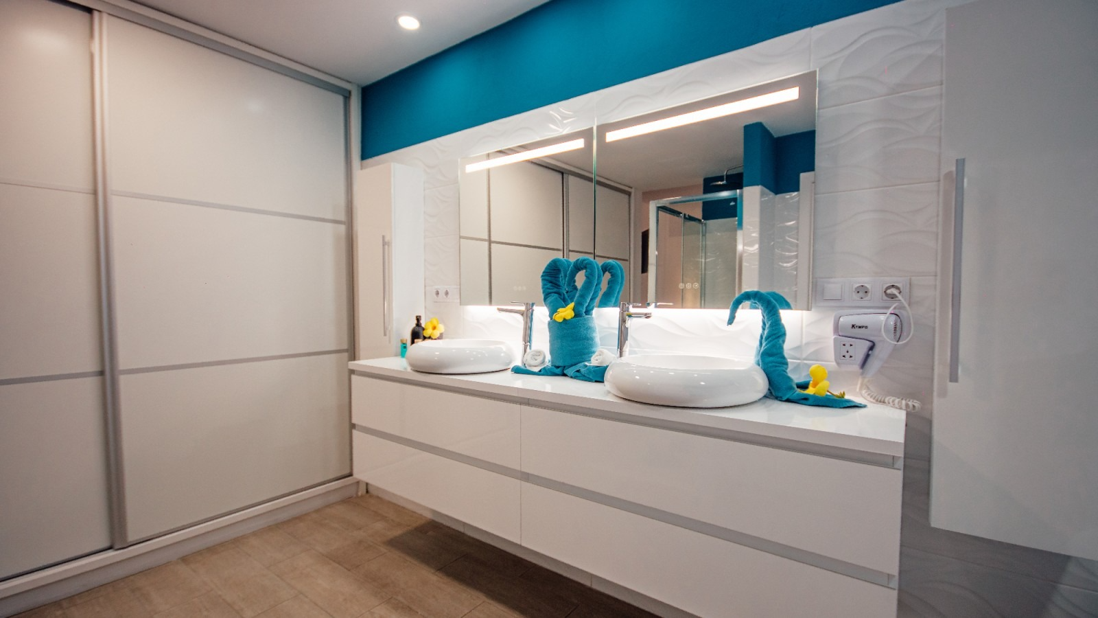 Modern bathroom vanity with white drawers, teal accents, mirrors, and neatly folded towels