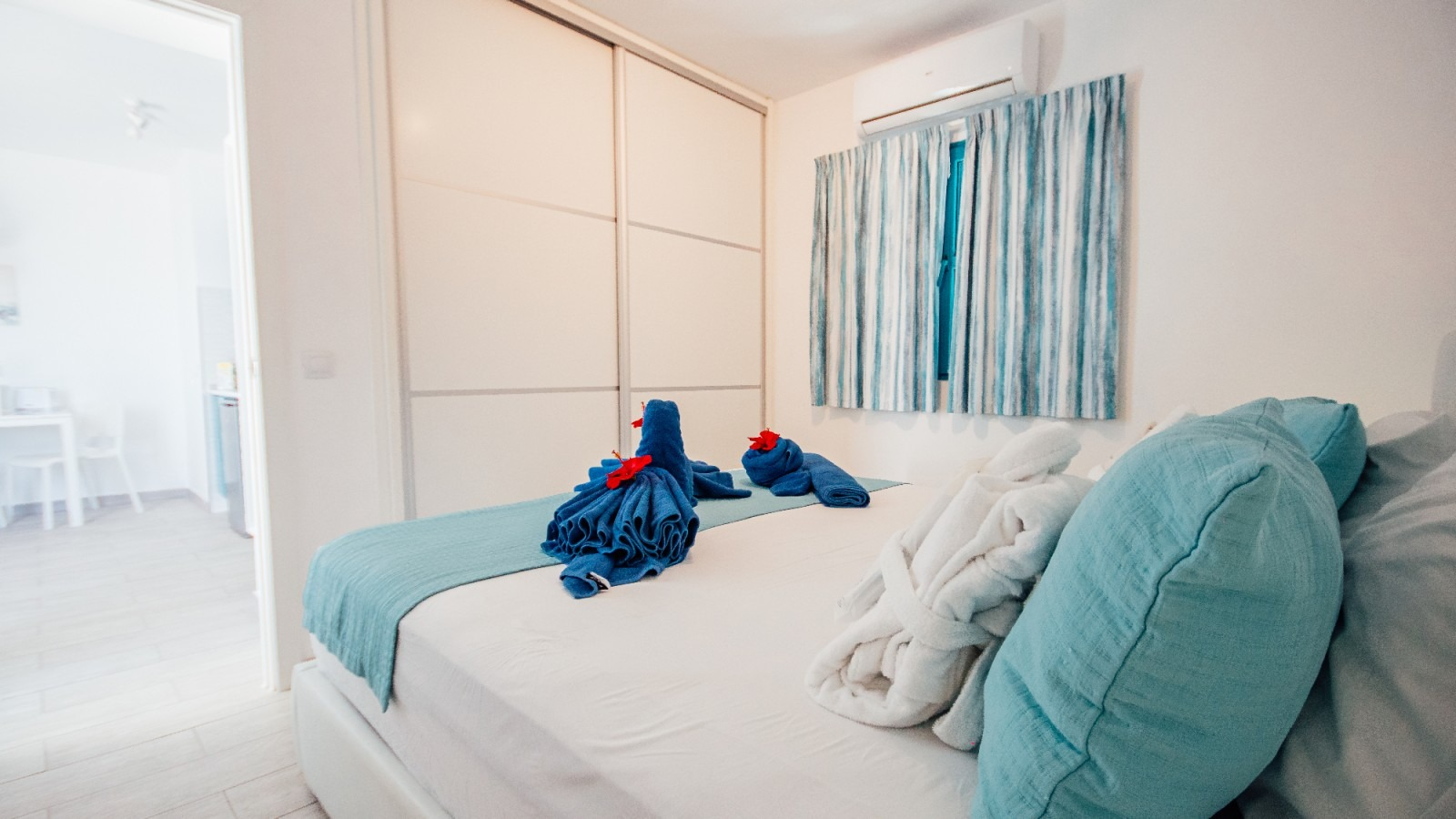 Bright bedroom with a white bed, teal pillows and curtains, and two stuffed toys on the bed.