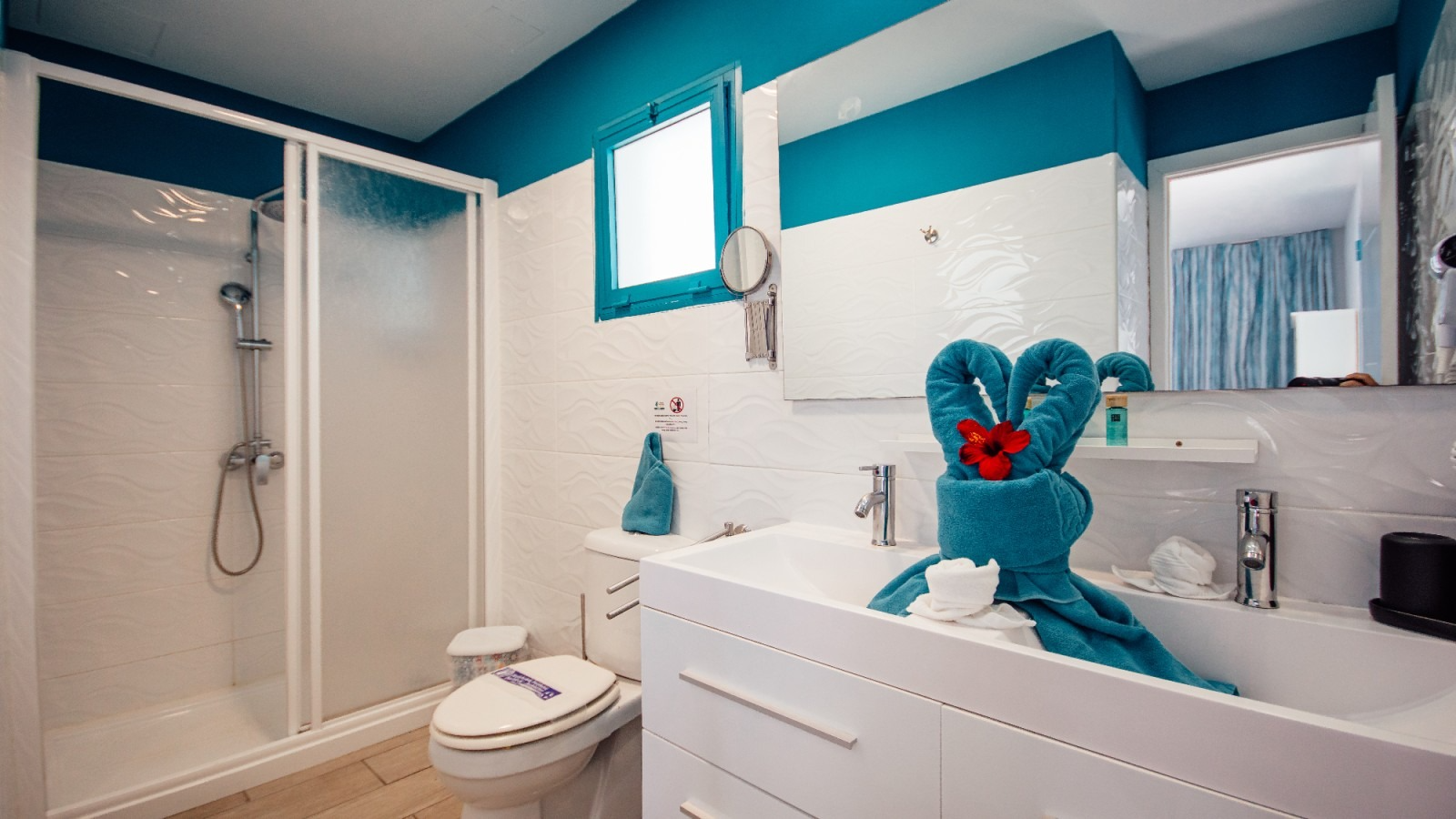Bright turquoise bathroom with a shower, toilet, vanity, and a blue bunny towel figure on the sink